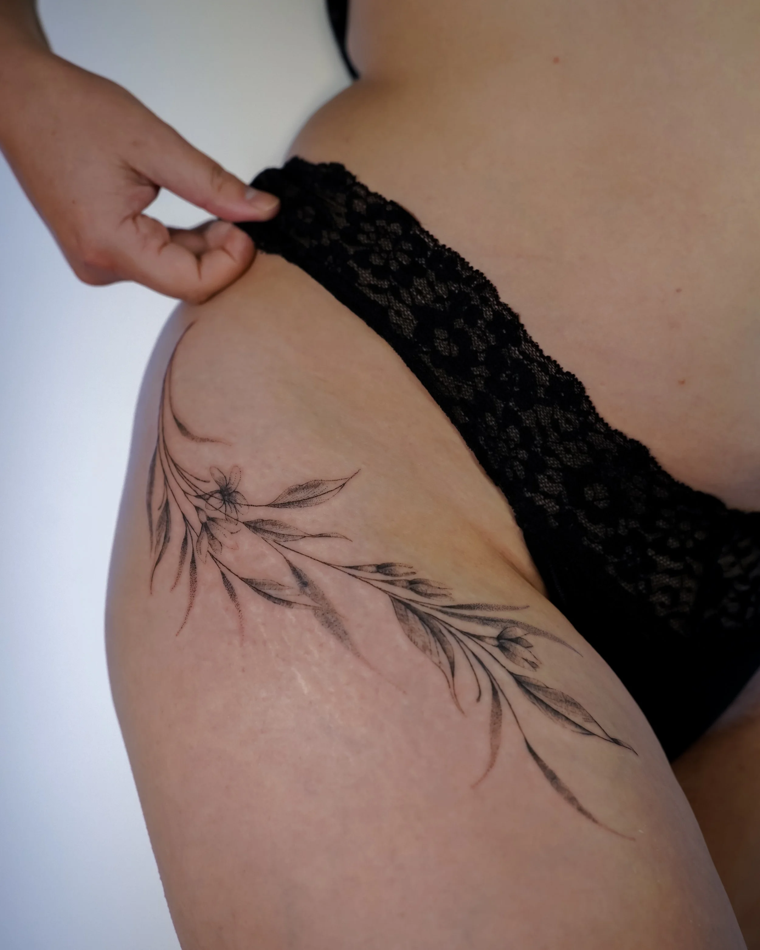 A close-up of a woman's thigh showing a black floral tattoo with leaves and flowers, partially hidden under a black lace stocking.