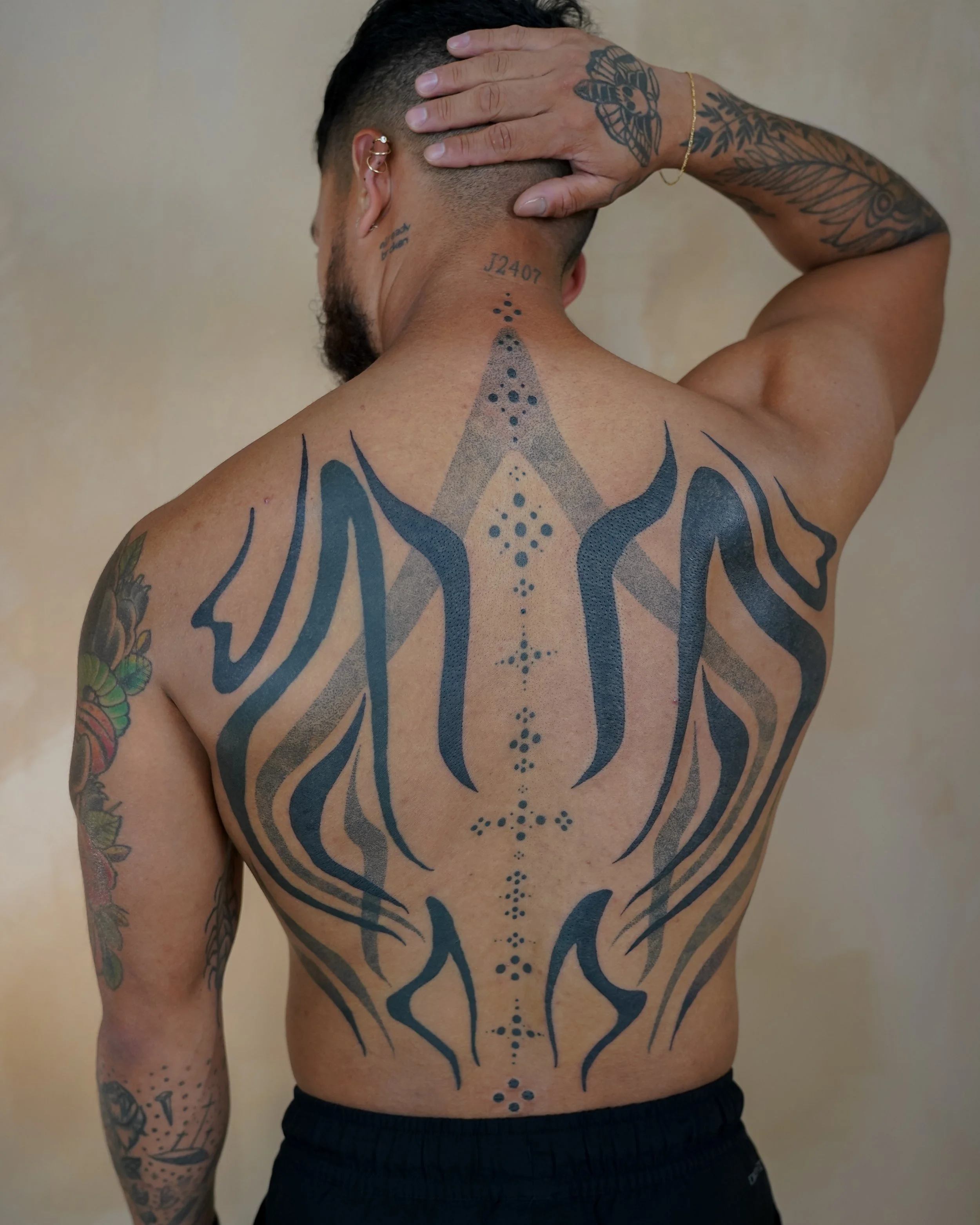 Back of a man with multiple tattoos, including a large tribal design on his back, a tattoo on his neck, and tattoos on his arms. He has short hair and is touching the back of his head with one hand.