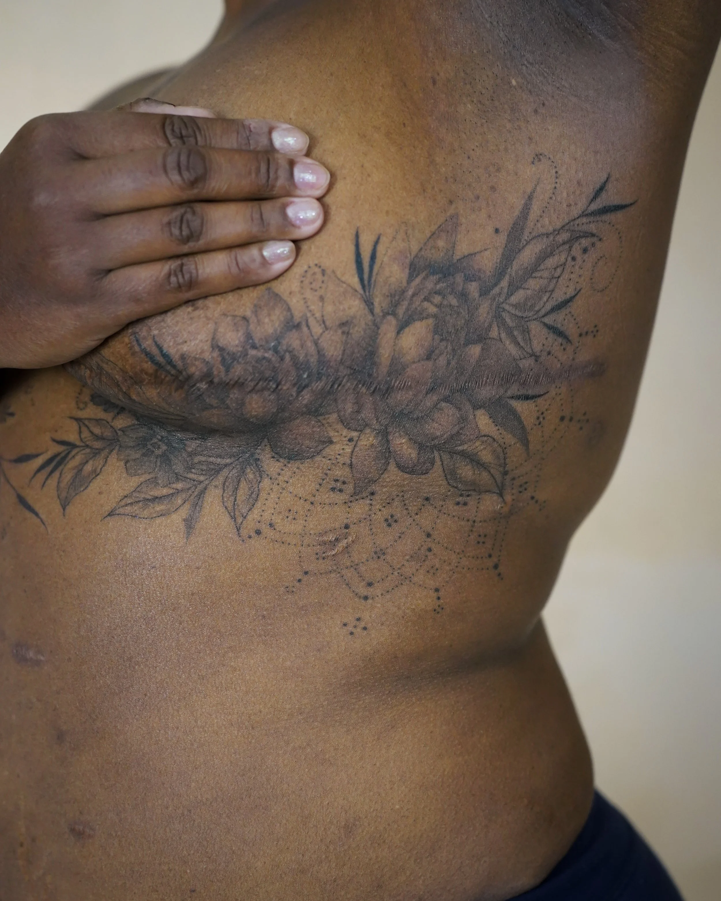 Close-up of a person's upper body with a floral tattoo on the chest, hand covering the chest, skin tone dark, background plain.