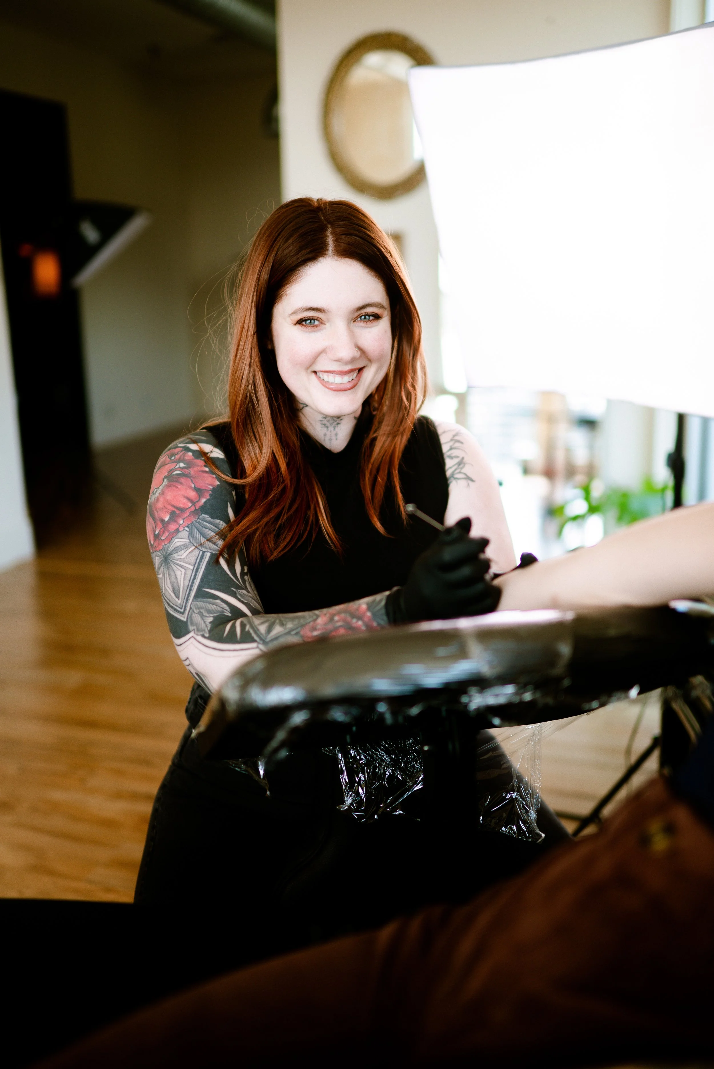 A woman with long red hair and tattoos on her arm smiling while tattooing.