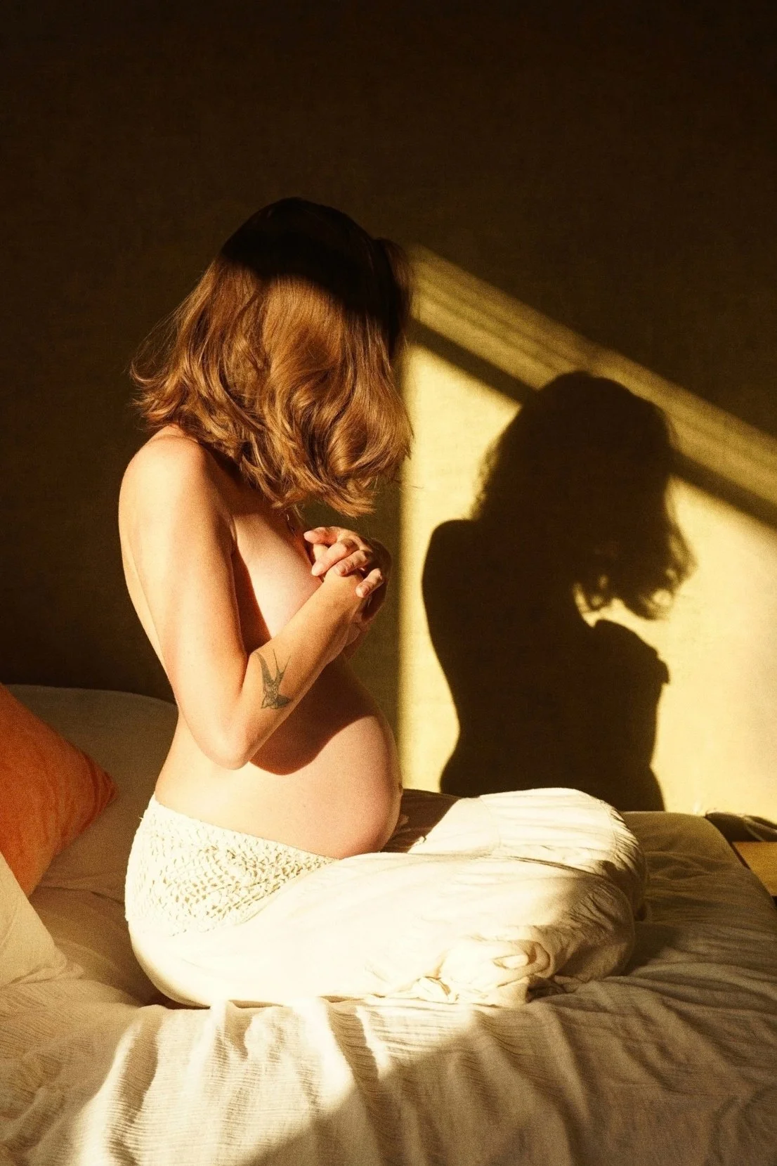 Zwangerschapsfotografie thuis: zwangere vrouw op bed in warm zonlicht, met haar schaduw op de muur