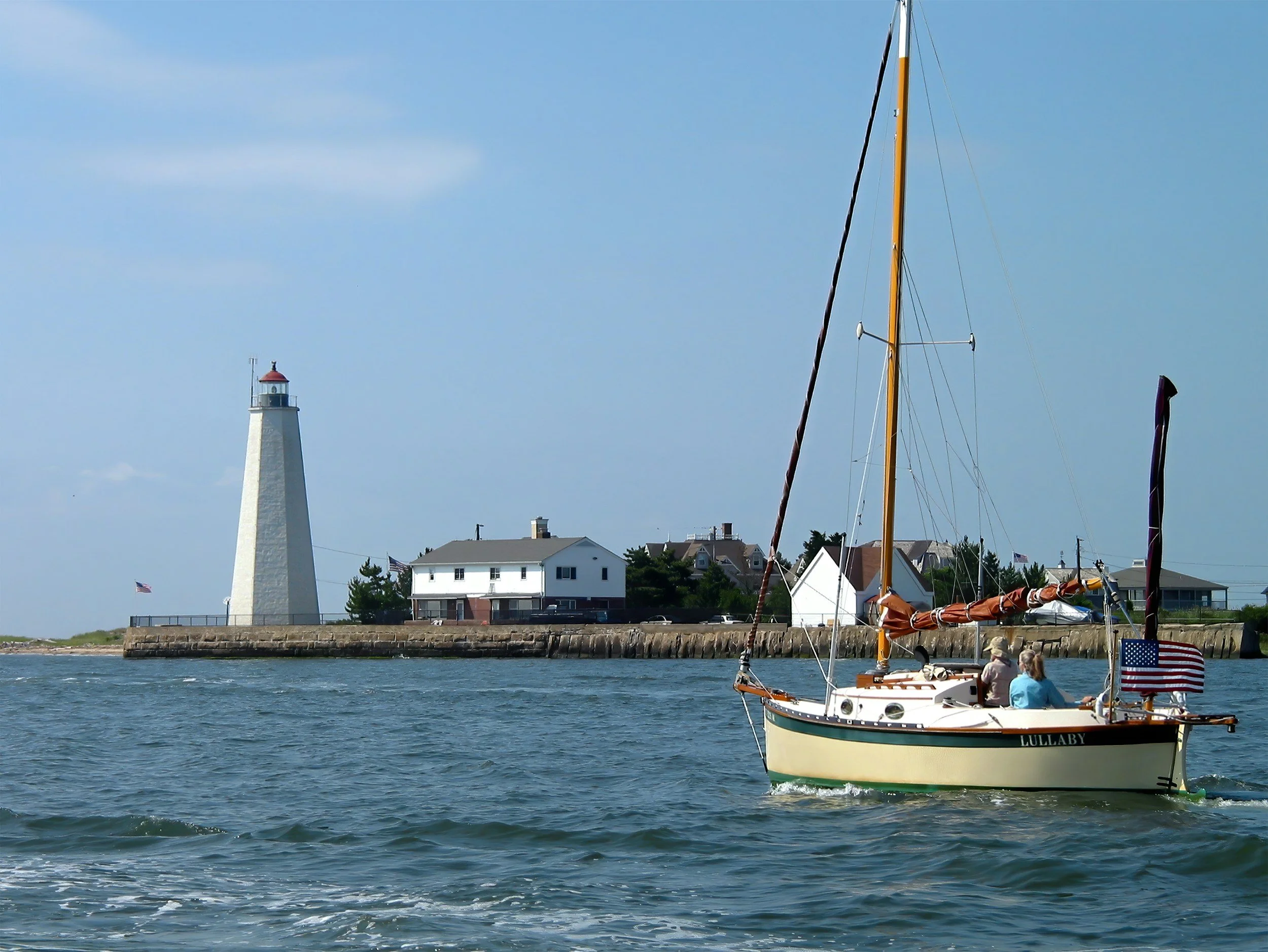 Sailboat on water near a lighthouse and shoreline houses under a clear blue sky.