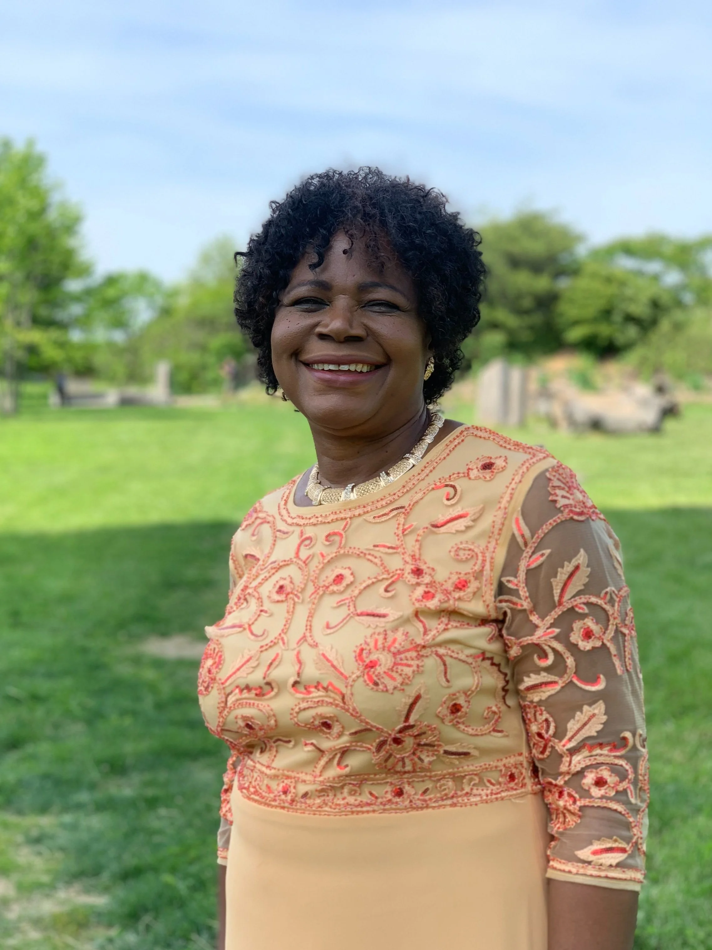 Smiling woman in an embroidered dress standing outside in a park with greenery in the background.