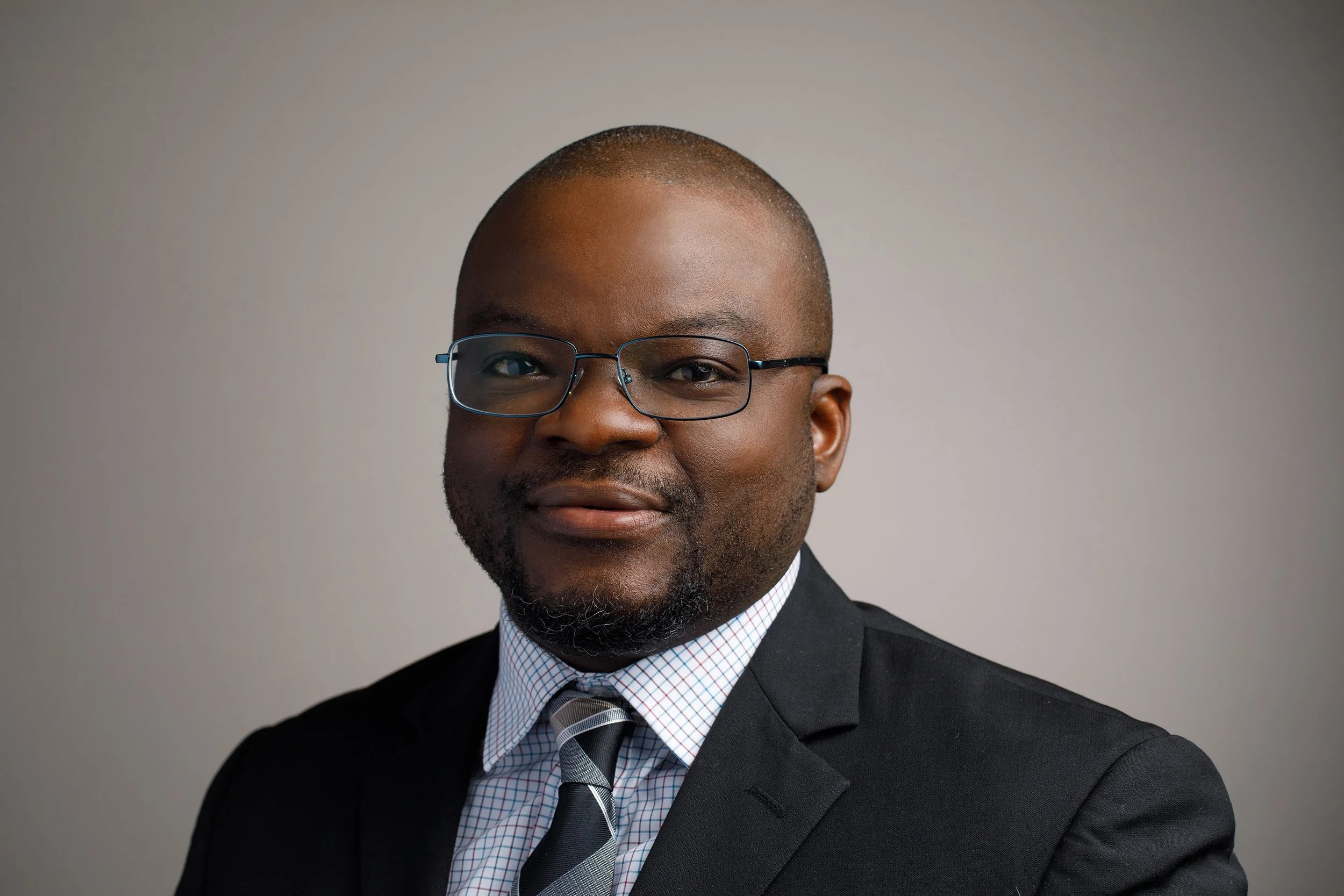 A man wearing glasses and a suit with a tie, smiling against a gray background.