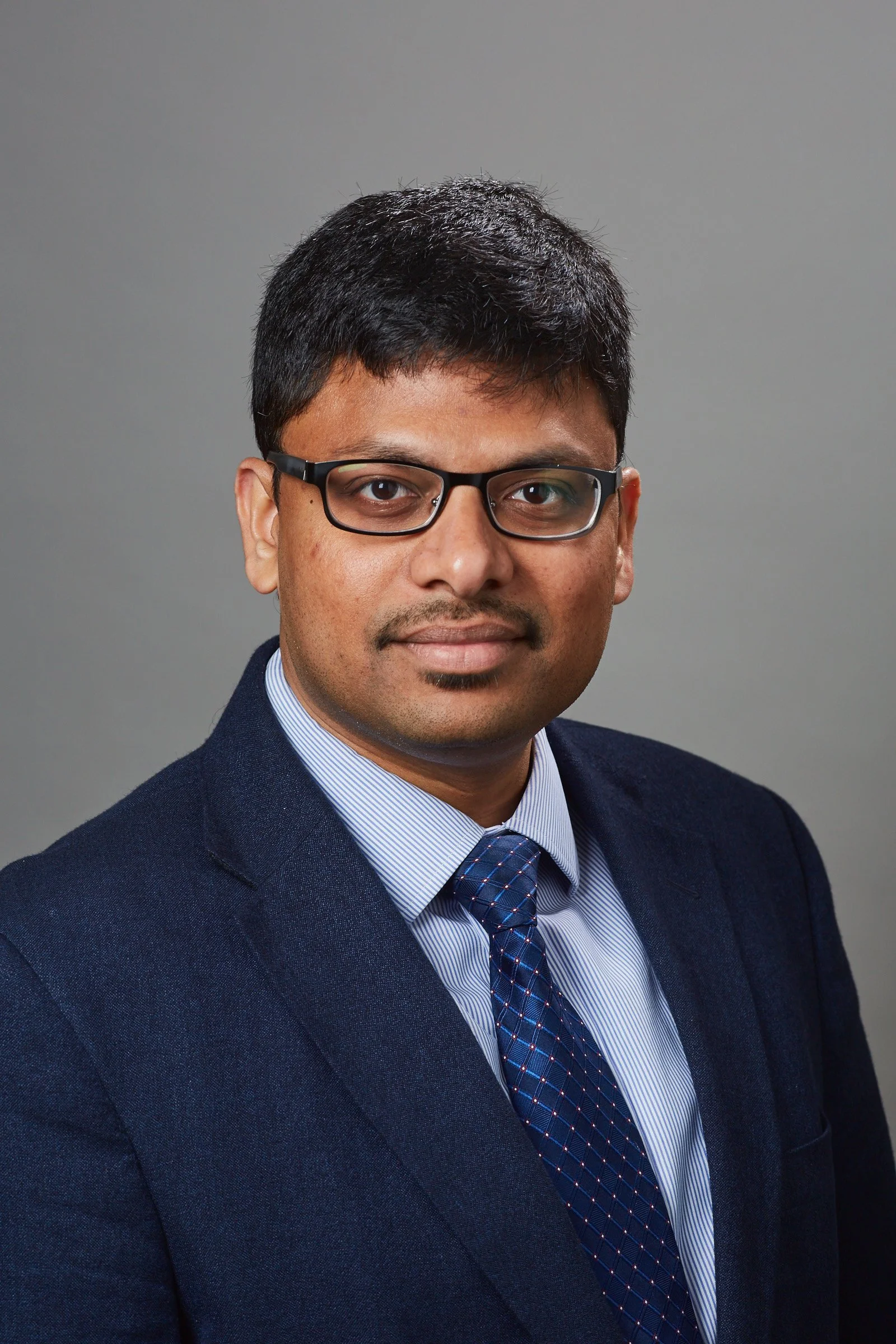 A man wearing glasses and a suit with a blue tie, posing for a professional portrait against a gray background.