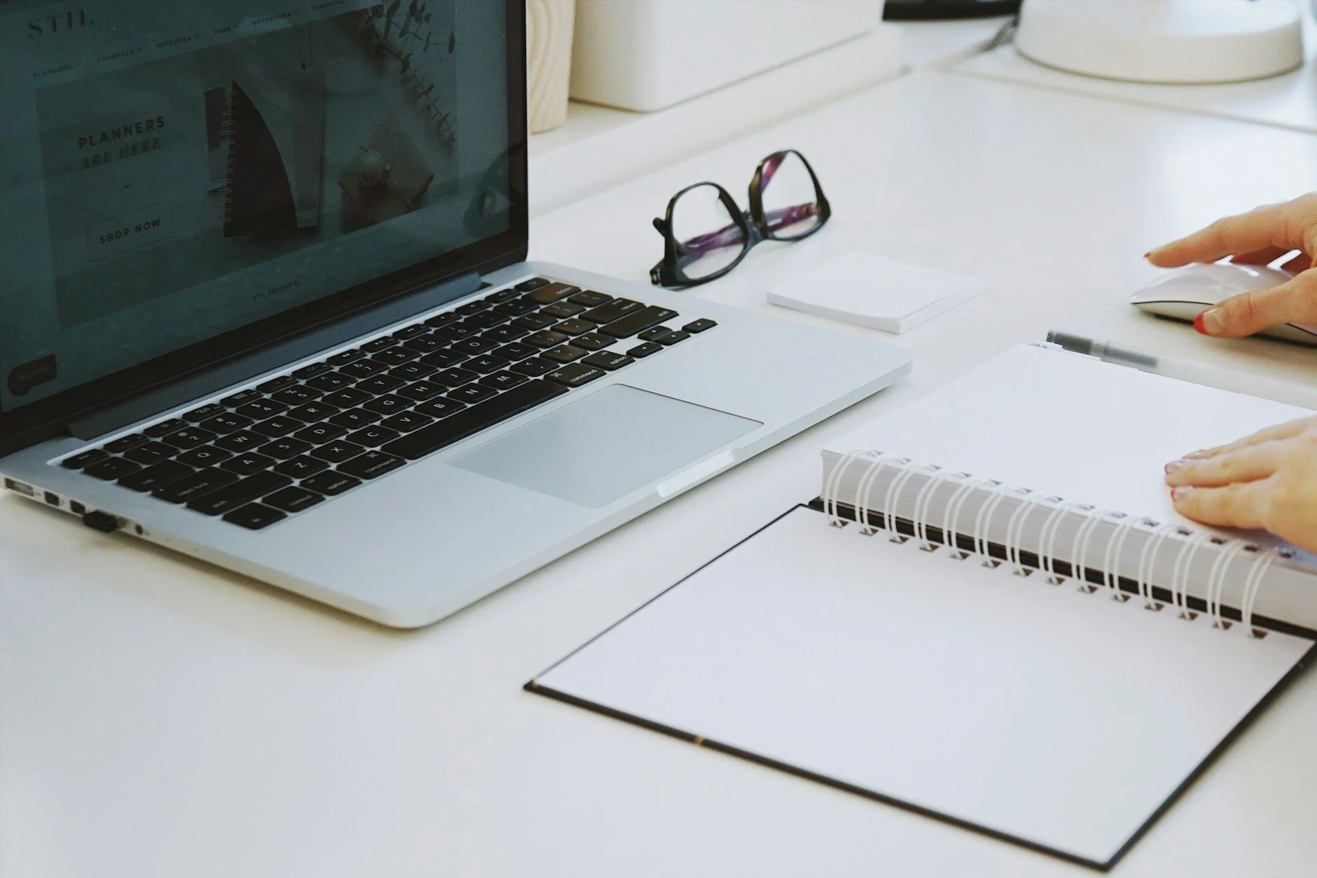 Laptop on a white desk with open notebook, glasses, sticky notes, and a person using a mouse.