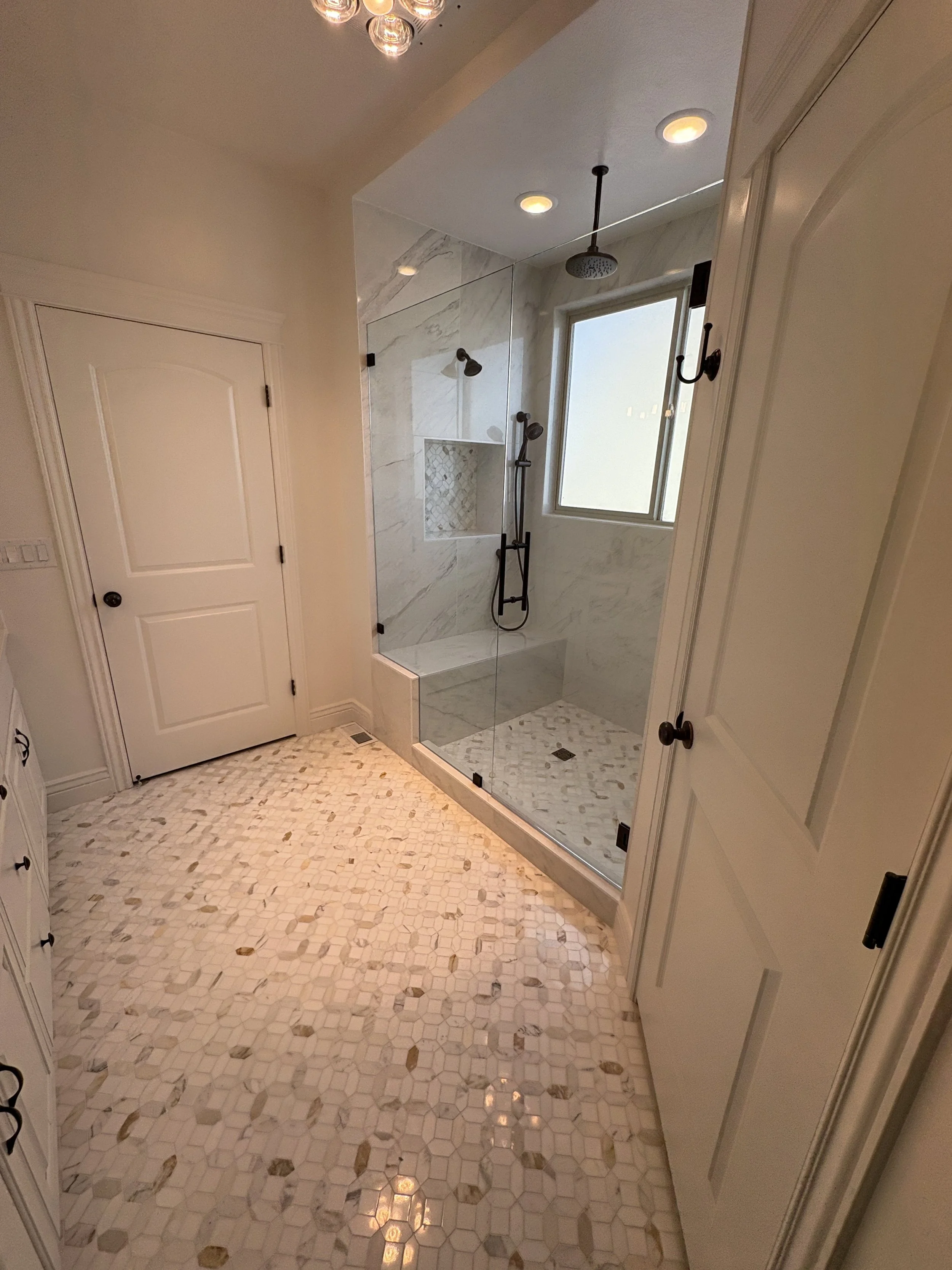 A bathroom with a tiled hexagonal floor, white cabinets, and a glass shower enclosure with marble walls, a window, and a rain shower head. Utah valley custom home builder saratoga springs