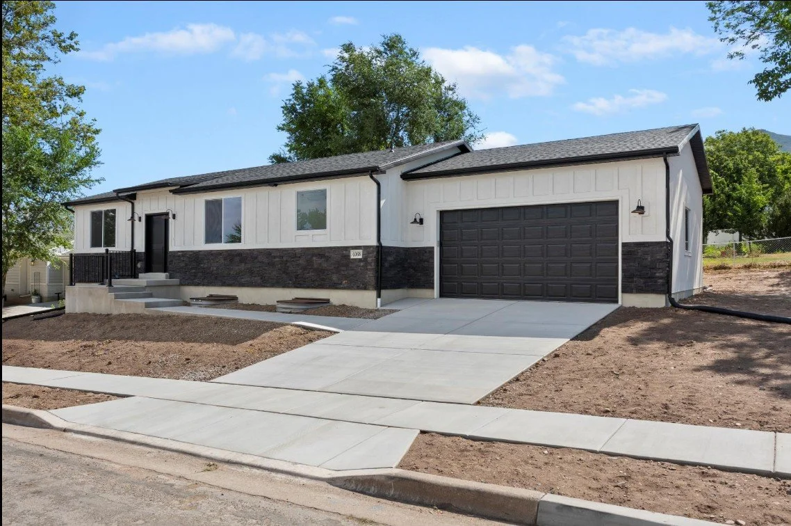 Newly constructed modern single-story house with black front door, attached garage, white siding, black accents, and a concrete driveway, surrounded by dirt yard and trees. Weber county, North Ogden, Utah