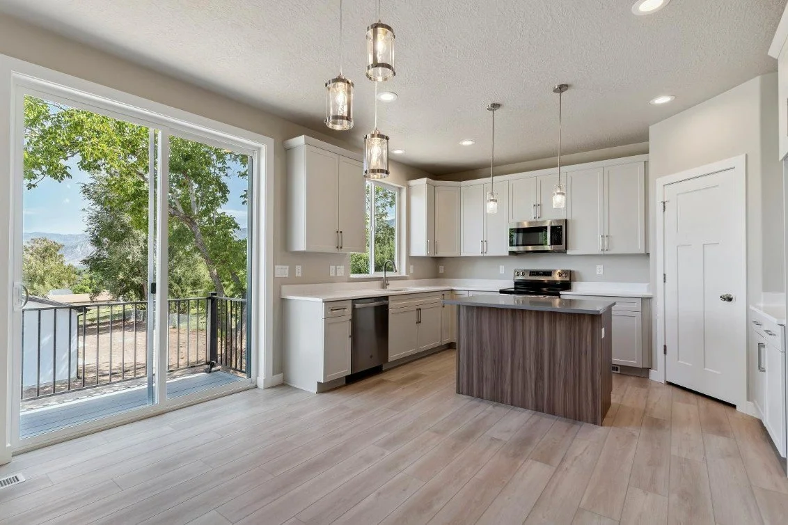 Modern kitchen with white cabinets, a small island, stainless steel appliances, pendant lights, a large window, and a sliding glass door leading to a patio with trees outside. Pleasant view utah