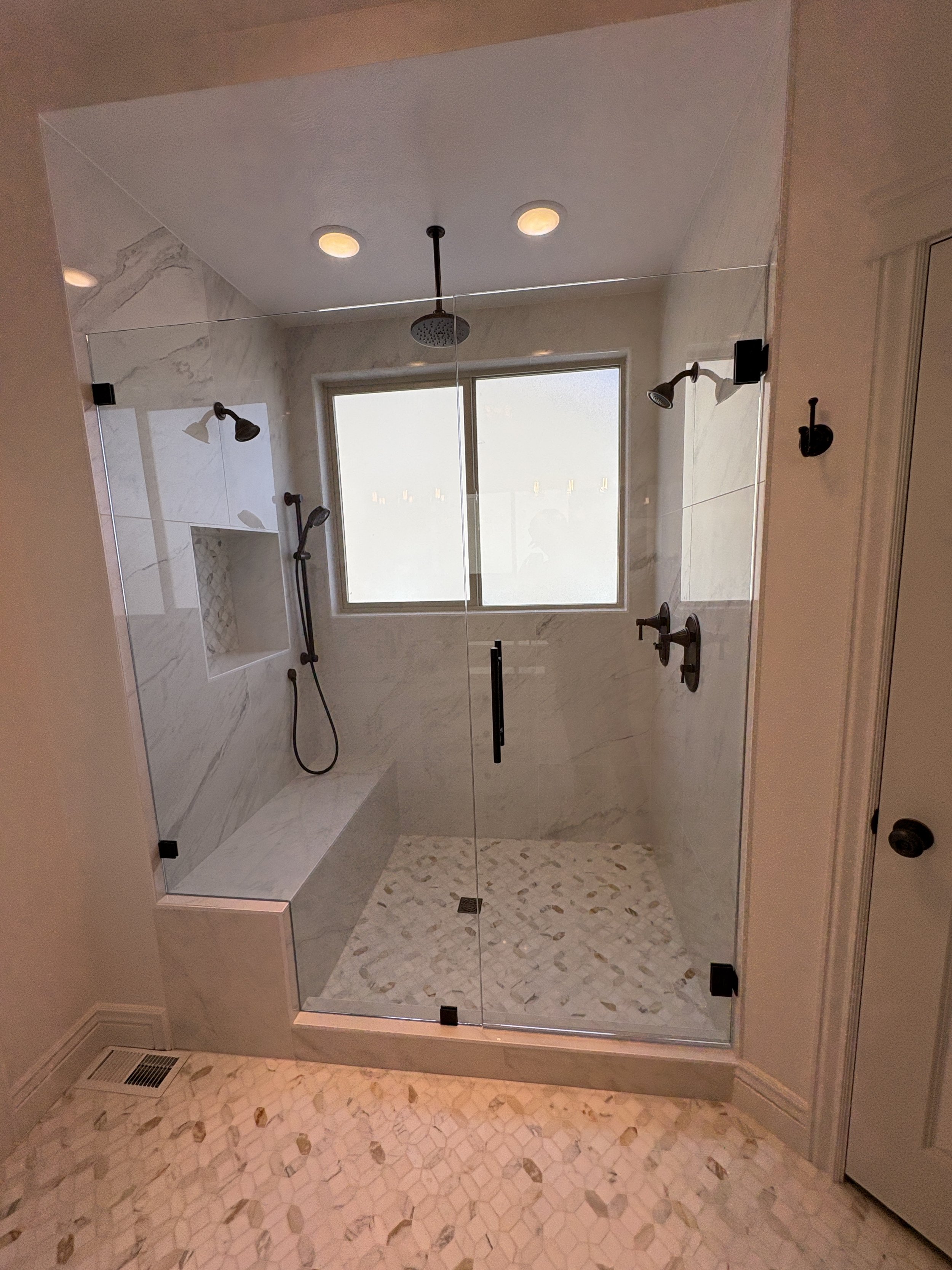A modern walk-in shower with a glass door, marble tiles on the walls, a window, and multiple showerheads including a rain shower and handheld wand, with pebble-style tiles on the floor. custom home remodeling construction, utah county lehi, alpine