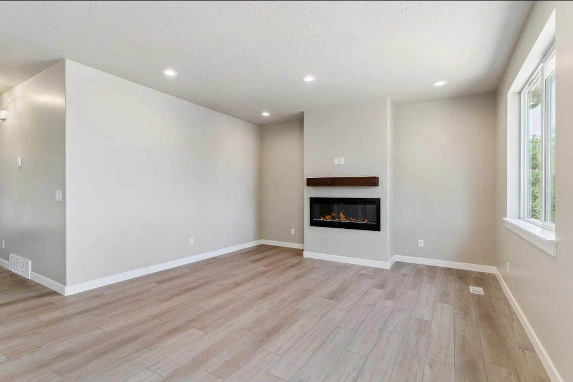 Empty living room with beige walls, wooden flooring, a corner fireplace with a wooden mantel, and a window letting in natural light. South Weber Utah