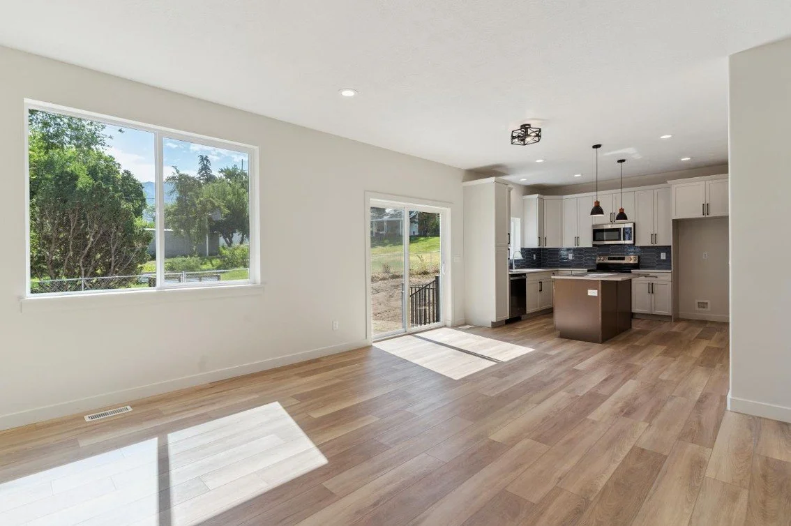 Empty living room with large window and sliding glass door leading outside, kitchen in background with white cabinets, black backsplash, and stainless steel appliances, wood flooring, sunlight streaming in. custom home builder Mountain green utah