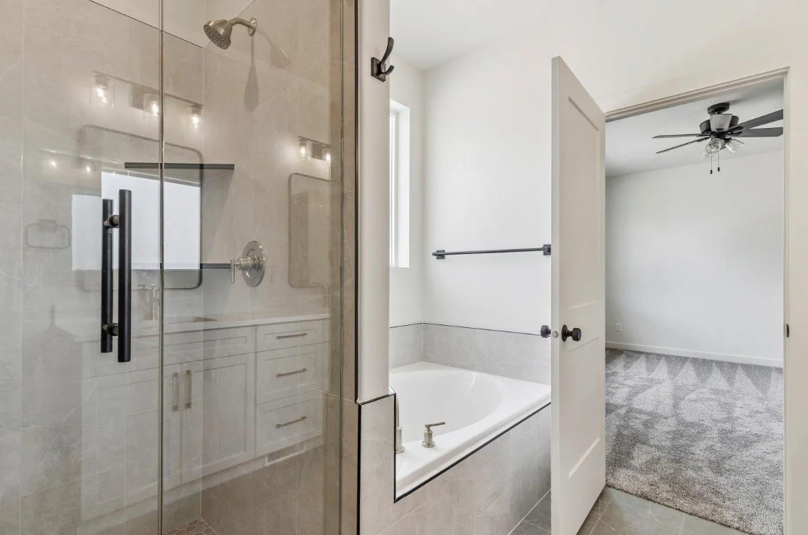 Bathroom with a glass shower enclosure on the left, a bathtub in the middle, and a doorway leading to a carpeted room with a ceiling fan in the background. ogden valley custom home builder