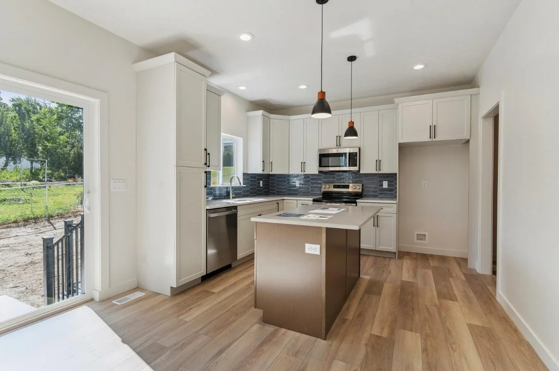 Modern kitchen with white cabinets, stainless steel appliances, gray backsplash, and a central island with a brown base. Large glass door leads outside, with wood flooring and pendant lighting. custom homes in morgan county utah