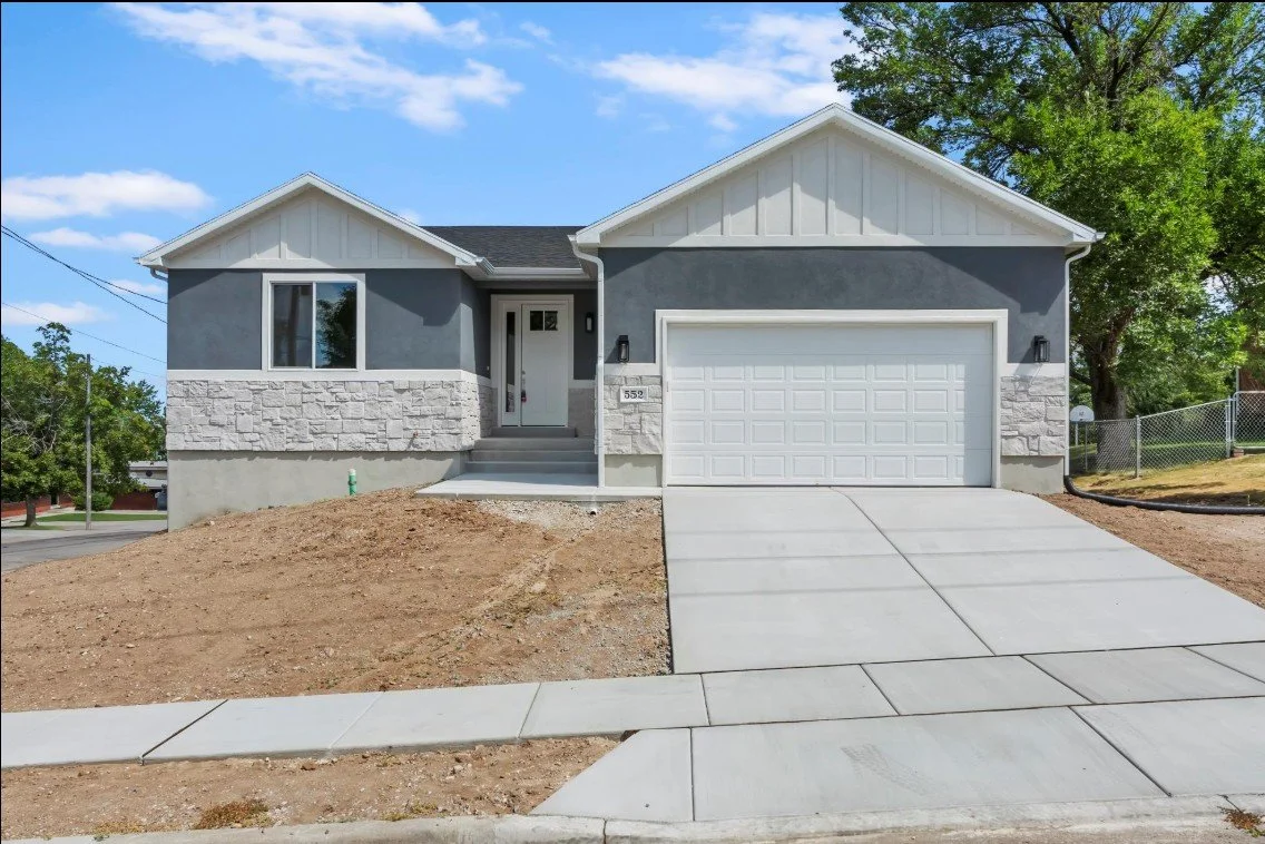 Newly built single-family house with gray exterior, white accents, a two-car garage, concrete driveway, front steps, and a front yard with soil, surrounded by a fenced backyard with trees. Weber county, South Ogden, utah