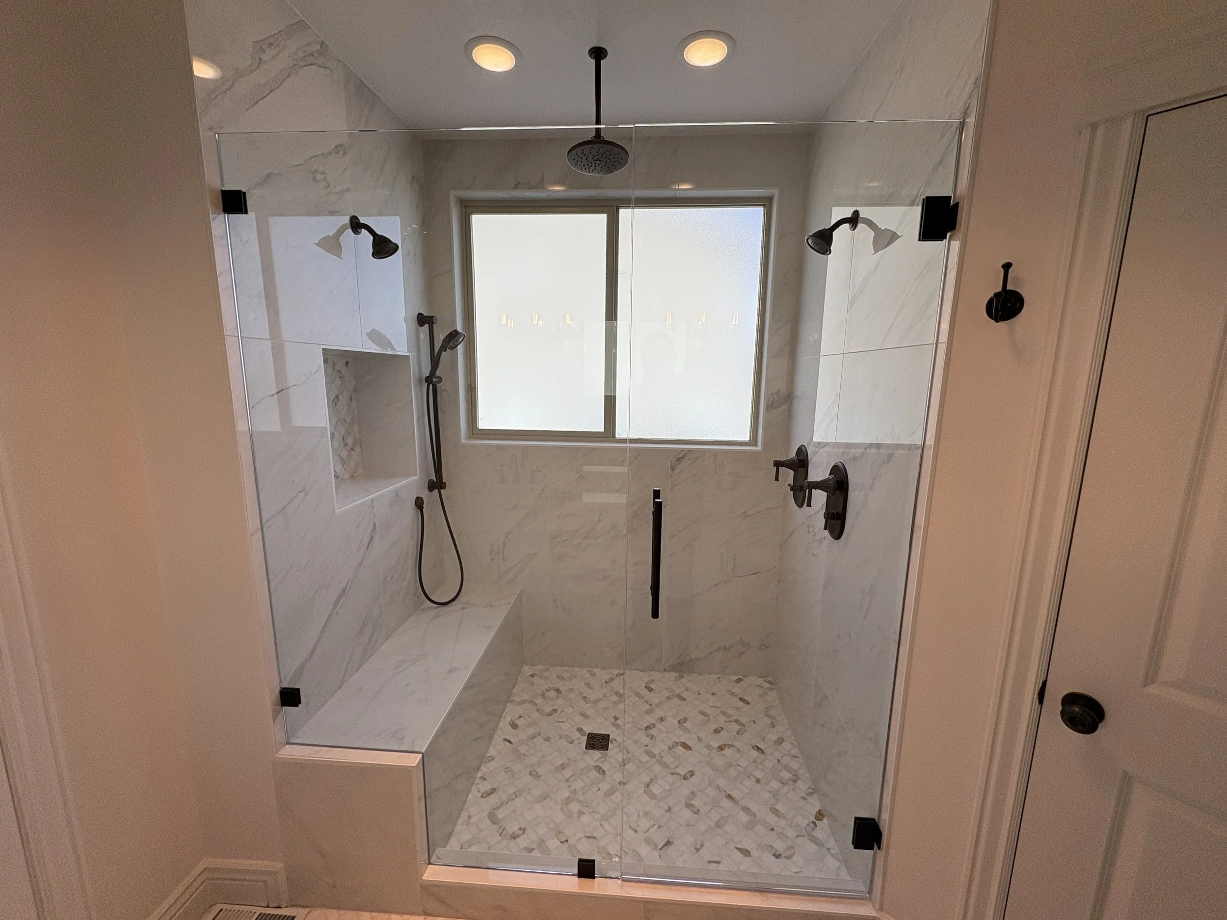 A walk-in shower with glass walls, marble tiles, two showerheads, and a window, in a modern bathroom. eagle mountain, cedar fort cutstom home builder