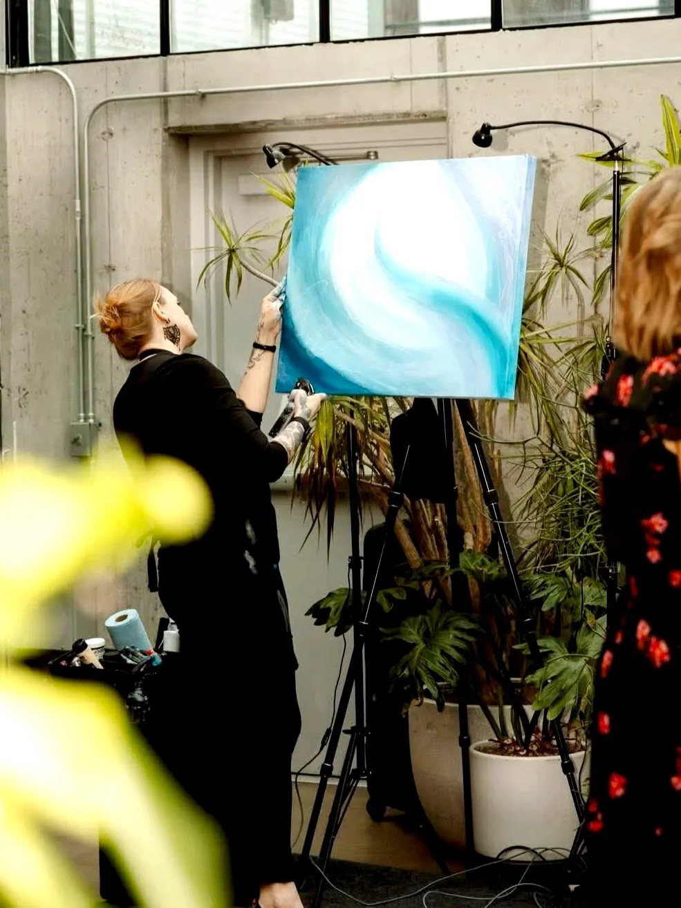 Hanna Bruer painting an abstract blue and white painting on a canvas set up on an easel, in a room with concrete walls and large windows, surrounded by potted plants during a wedding at the Tinsmith.