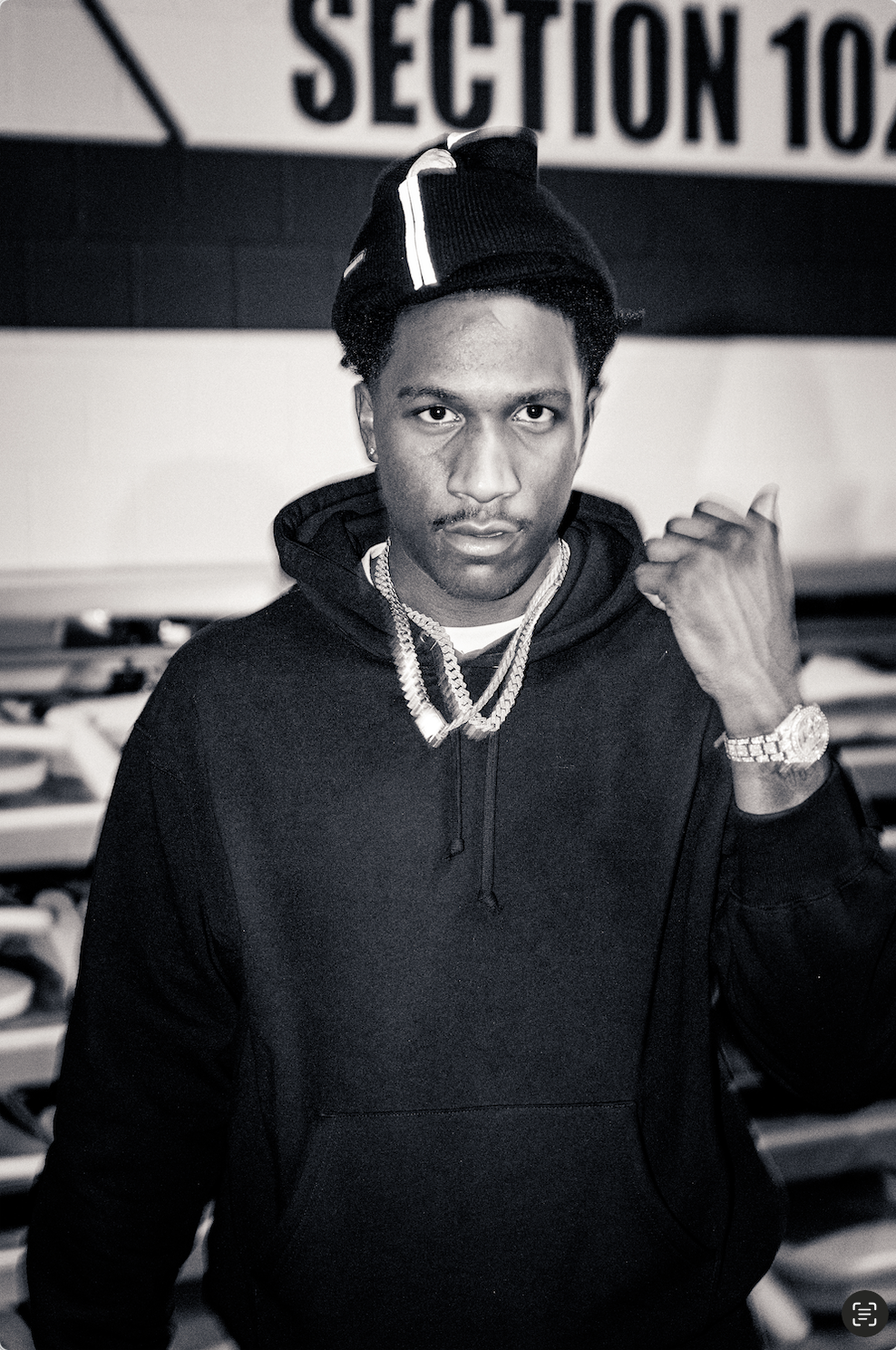 A young man wearing a black hoodie, beanie, and jewelry, posing indoors with a serious expression, with a sign that reads 'SECTION 102' in the background.