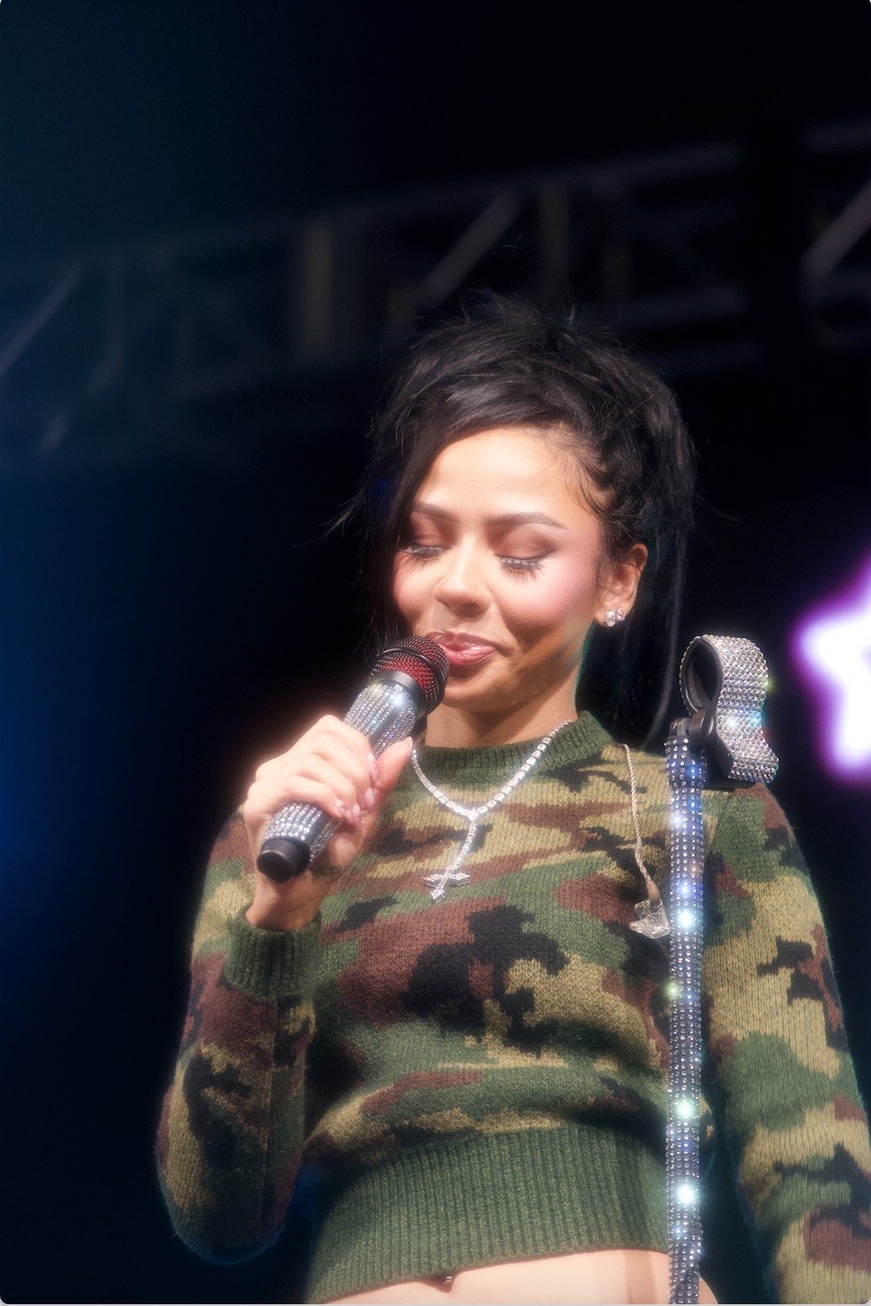 A woman with dark hair and closed eyes holds a microphone encrusted with rhinestones, standing on stage with dark background and bright lights.