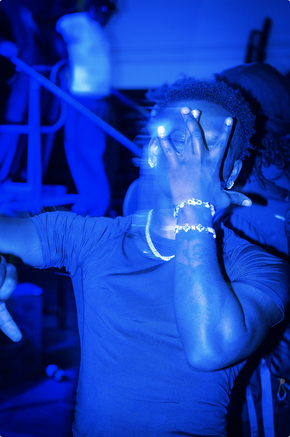 A person with curly hair, wearing jewelry, is illuminated by blue light, holding glasses up to their face with their hand, at a party or nightclub.