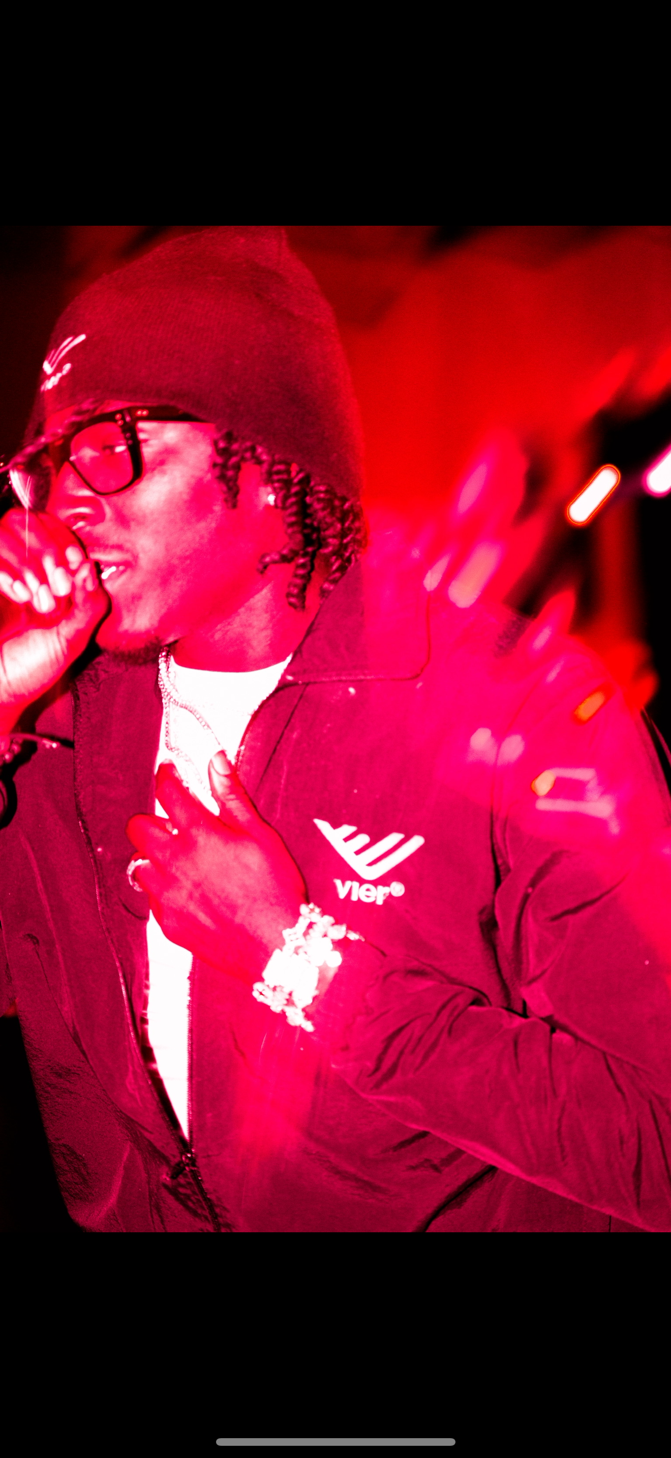 Person wearing glasses and a beanie, singing or speaking into a microphone, dressed in a jacket with a logo, with a red background and some bright lights.