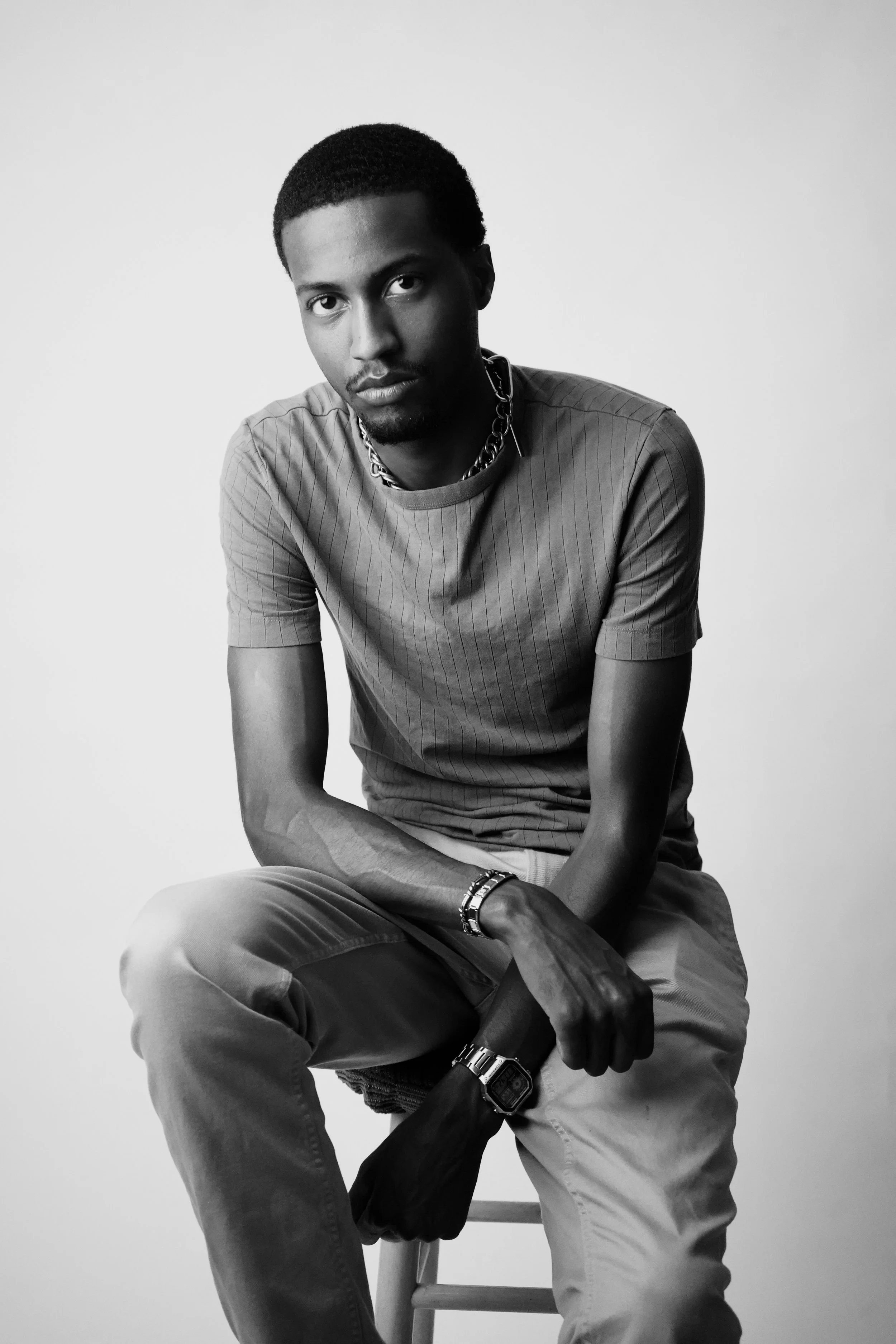 Black and white portrait of a young man sitting on a stool, wearing a short-sleeved shirt, pants, and accessories including a chain necklace, bracelet, and wristwatch, with a plain light background.