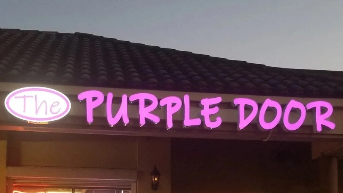 Neon sign reading 'The Purple Door' with a purple and pink color scheme, mounted on a building with a tiled roof.