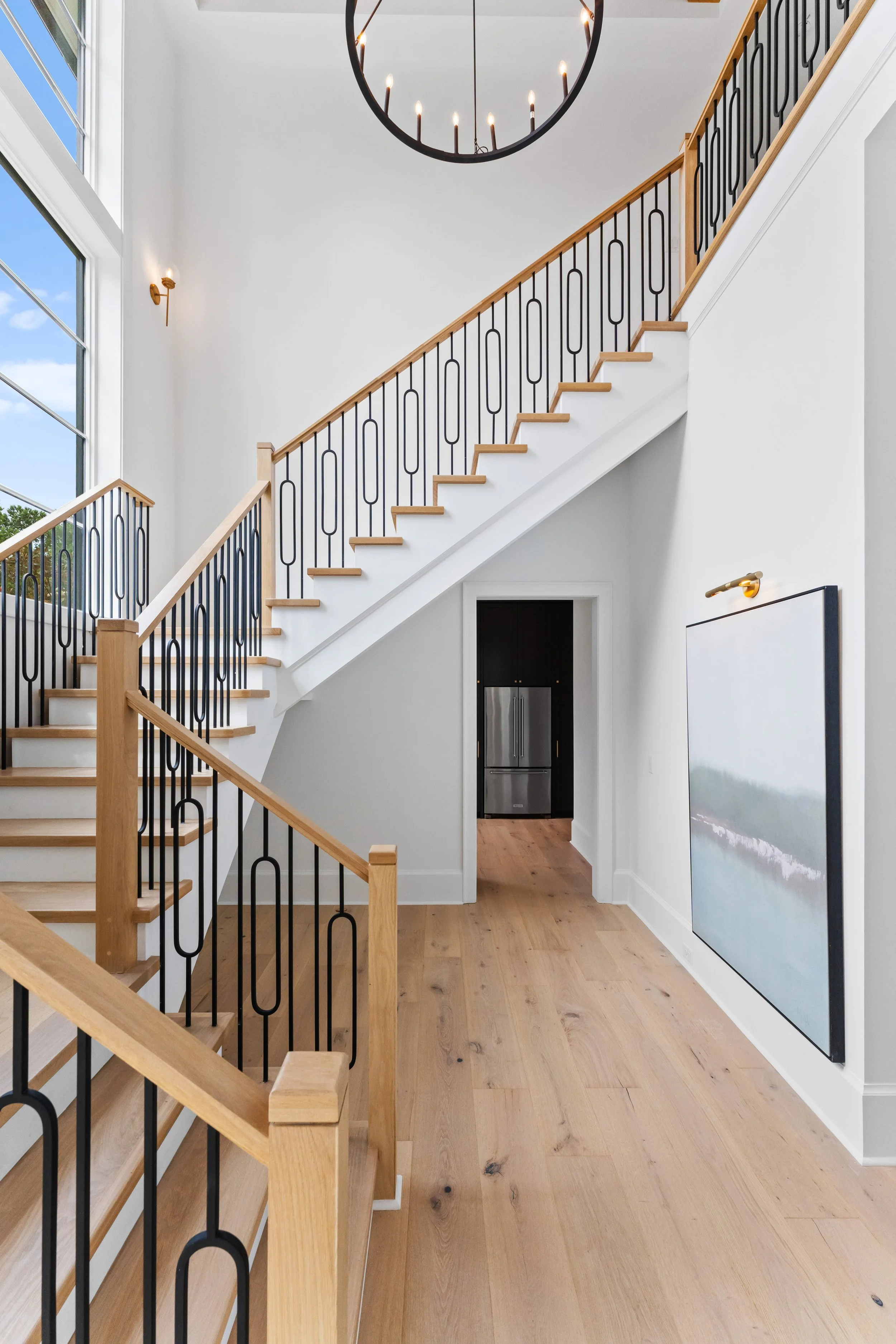 Interior view of a staircase with wooden handrails and black metal balusters, leading to an upper floor with large windows letting in natural light, a chandelier hanging from the ceiling, a wall-mounted light fixture, and a doorway revealing a kitche