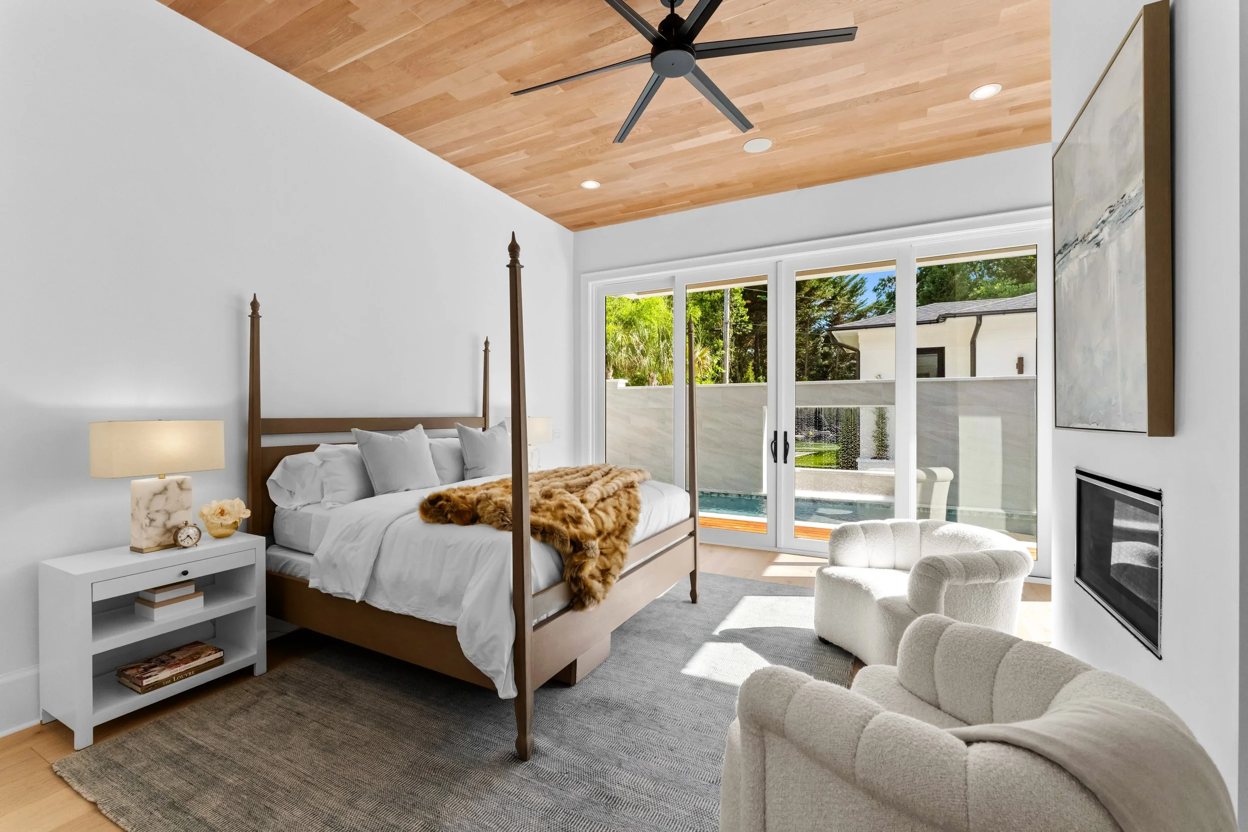 Bedroom with large sliding glass doors showing outdoor pool and patio, white walls, wooden ceiling, modern decor, and a four-poster bed with white bedding and fur throw.