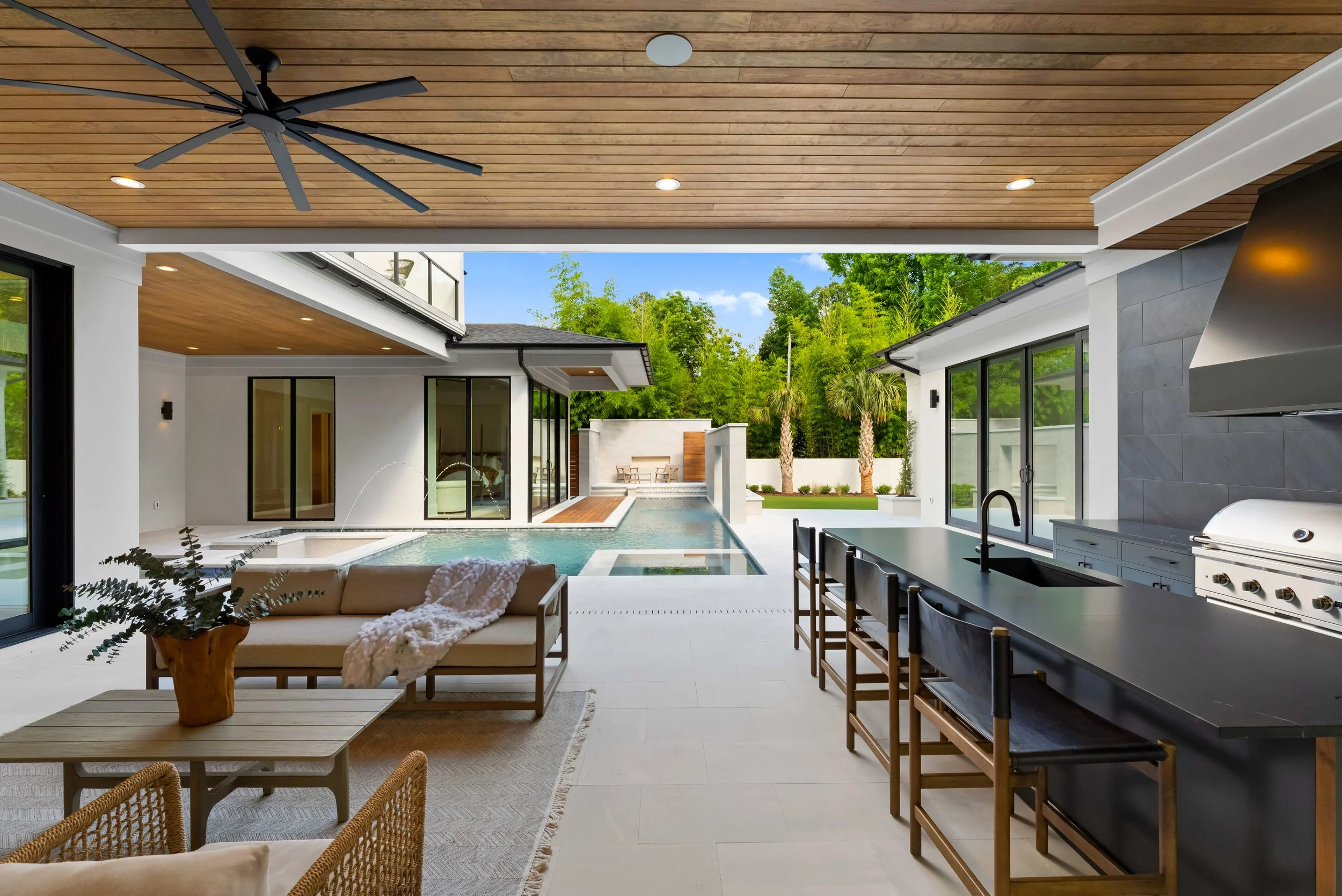Modern backyard with a pool, outdoor kitchen, and seating area under a wooden ceiling with ceiling fan, with lush green trees in the background.