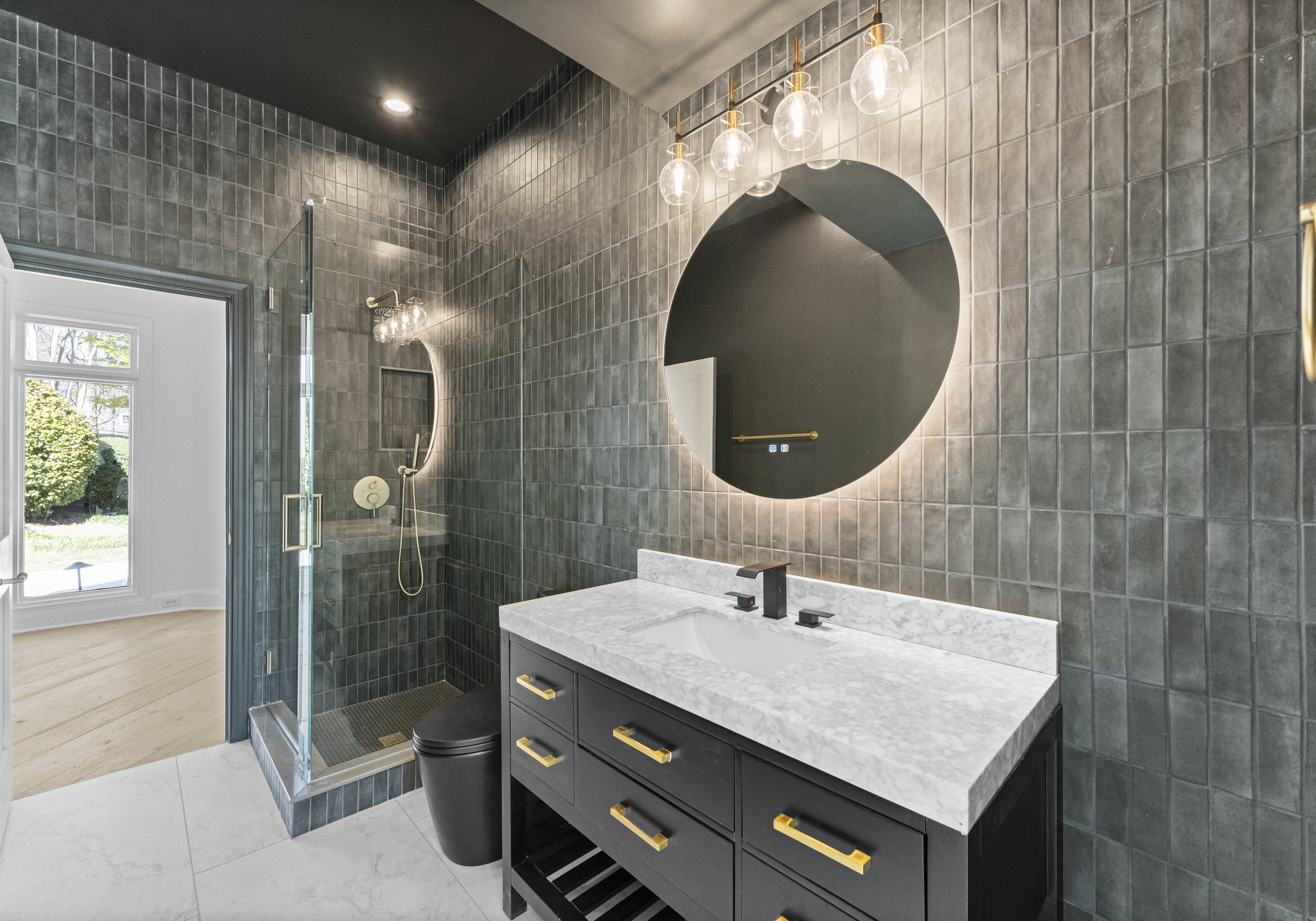 Modern bathroom with dark tiled walls, black vanity with gold handles, marble countertop, round mirror with backlight, and walk-in shower with glass door and black toilet.