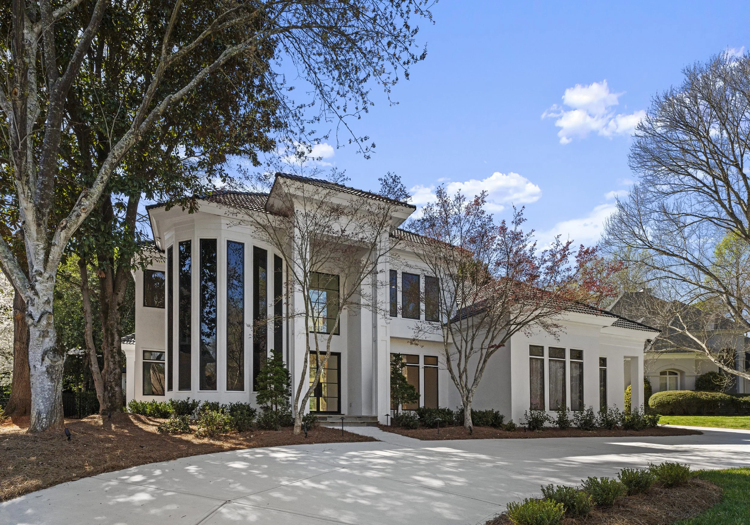 A modern white house with multiple large windows, surrounded by trees and a curved driveway, under a blue sky with some clouds.