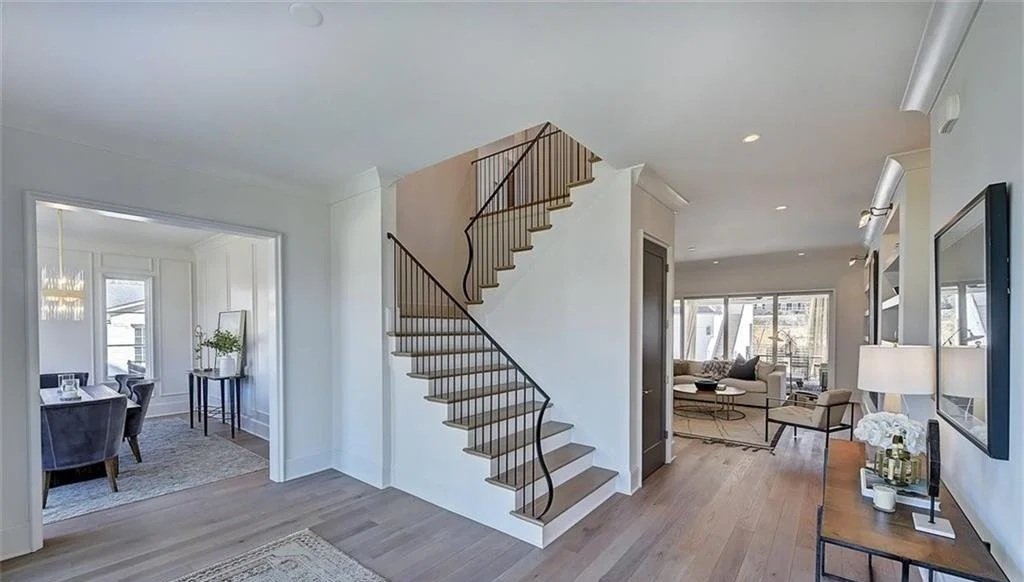 Interior view of a modern home showcasing a staircase with wooden steps and black railing, a dining area on the left, and a living room on the right with large windows and seating.