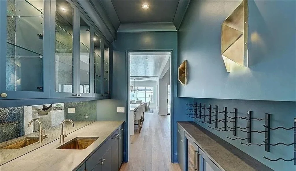 A modern kitchen area with blue cabinetry, a small double sink, and decorative wall sconces, leading to a dining area with large windows.