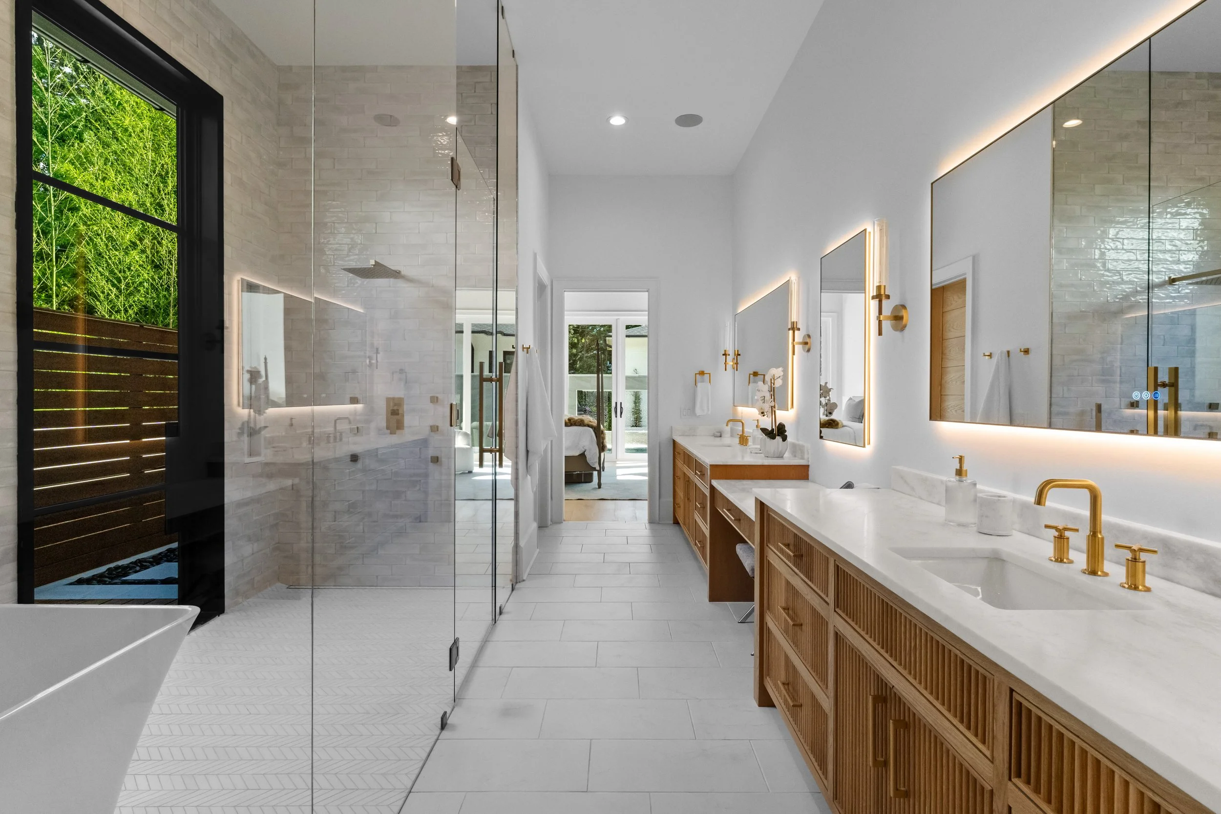 Modern bathroom with double vanity, gold fixtures, large mirrors, white marble countertops, wood cabinets, and a walk-in shower with glass doors, adjacent sliding glass doors leading to an outside area.