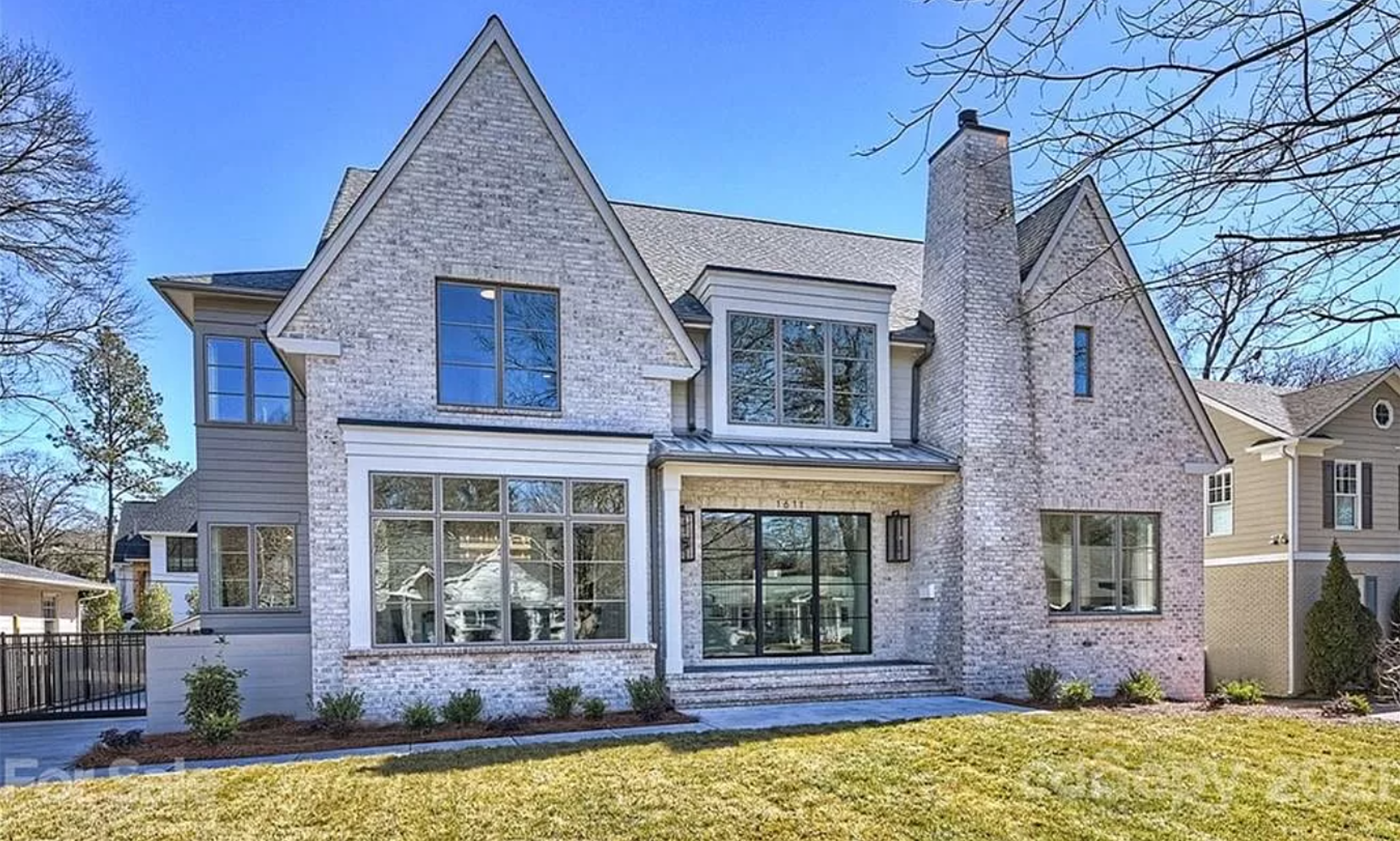 Modern two-story house with brick and siding exterior, large front windows, and a front porch, surrounded by a manicured lawn and leafless trees against a clear blue sky.