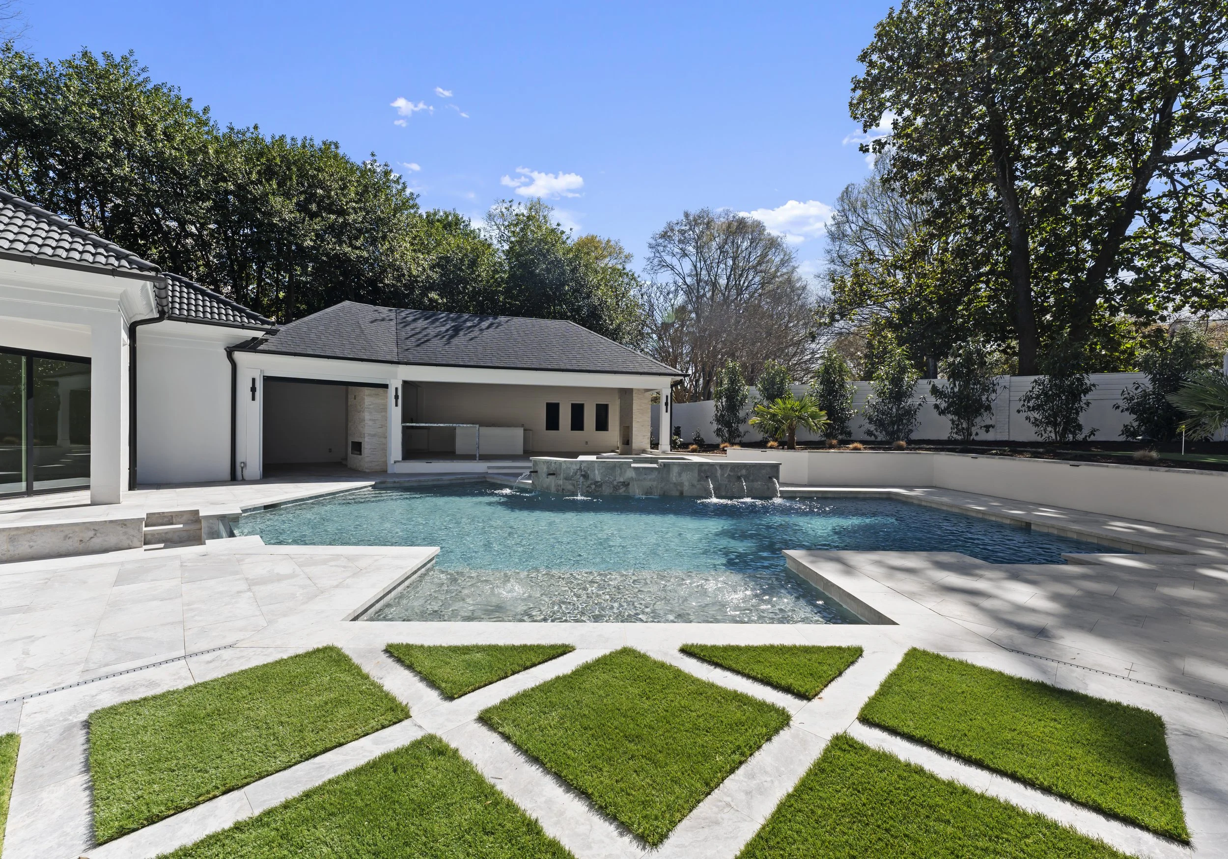 Modern backyard with a swimming pool, waterfall feature, and manicured lawn under a clear, sunny sky.
