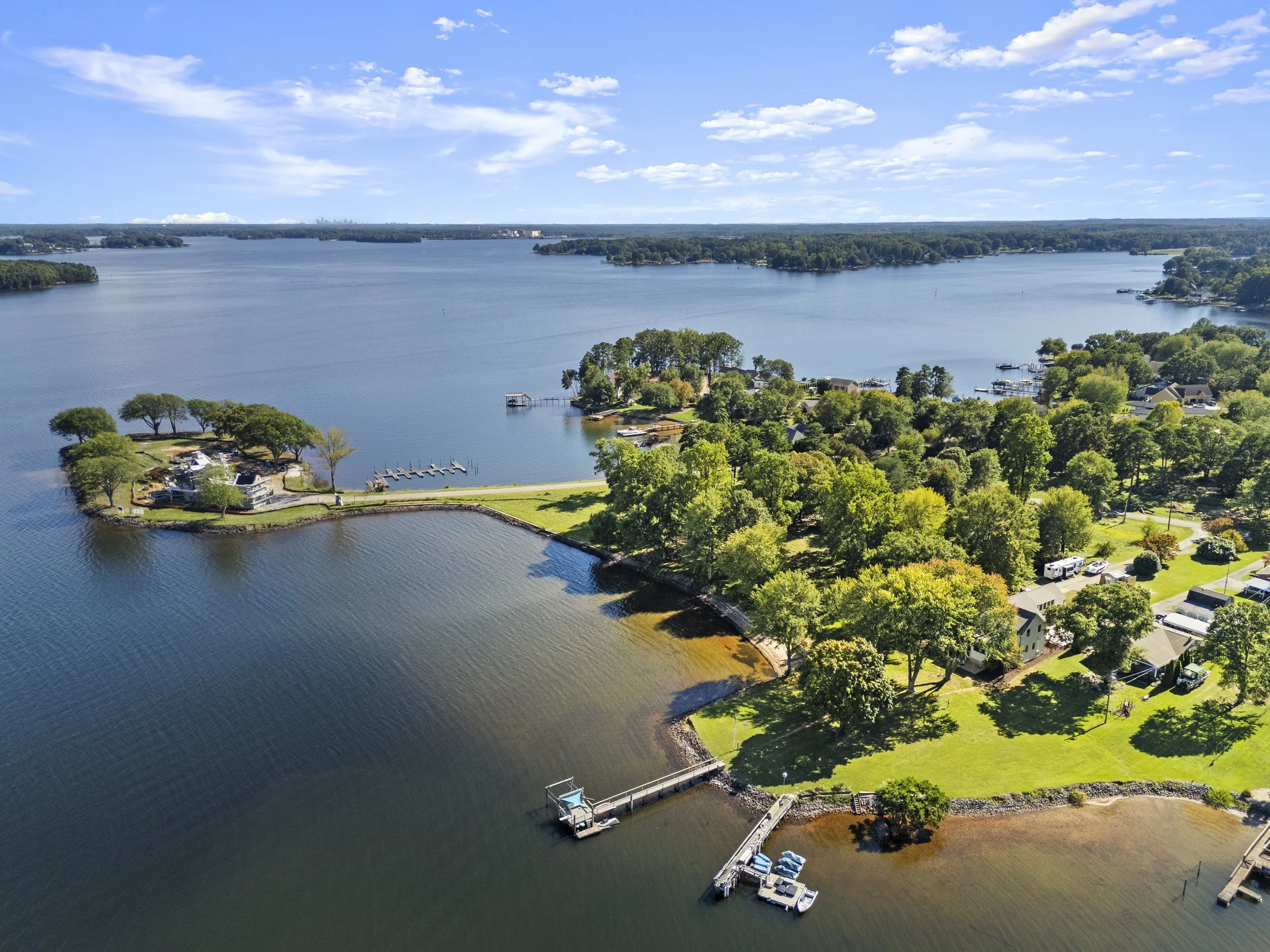 Aerial view of a lakeside community with green trees, houses, docks, and boats on a sunny day with a partly cloudy sky.