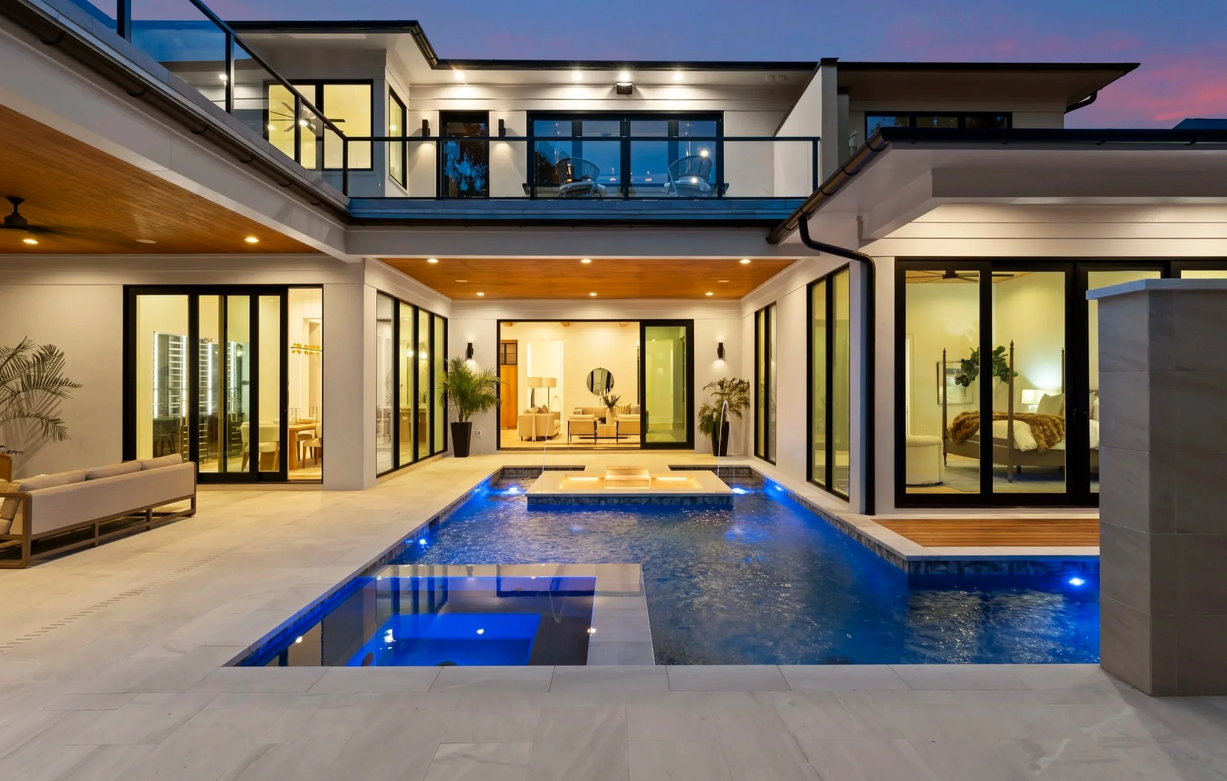 Modern house backyard with a swimming pool, outdoor seating, and large glass windows and doors showing interior rooms, during dusk.