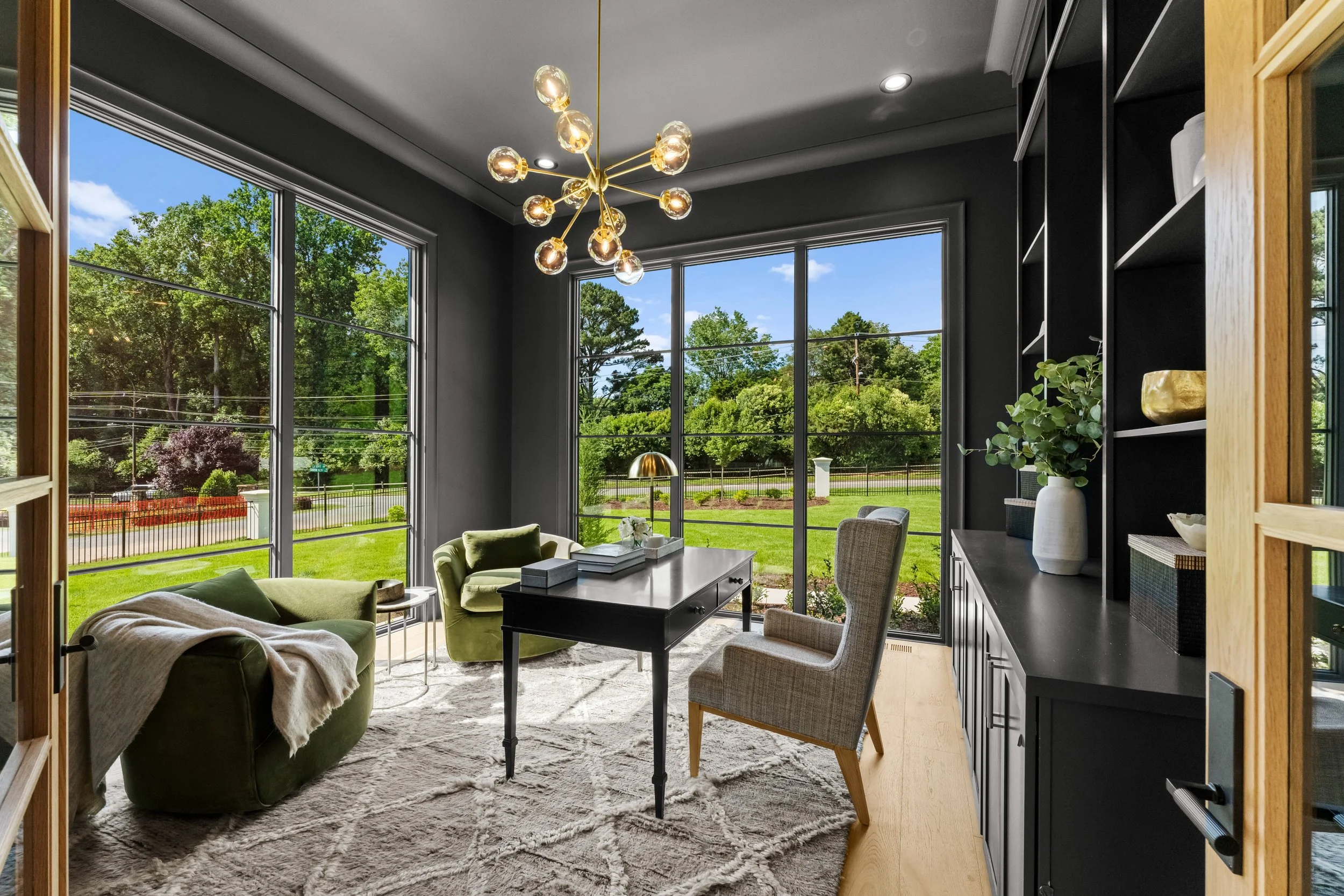 Modern living room with large windows, green and beige furniture, black wall with shelves, and a gold chandelier.