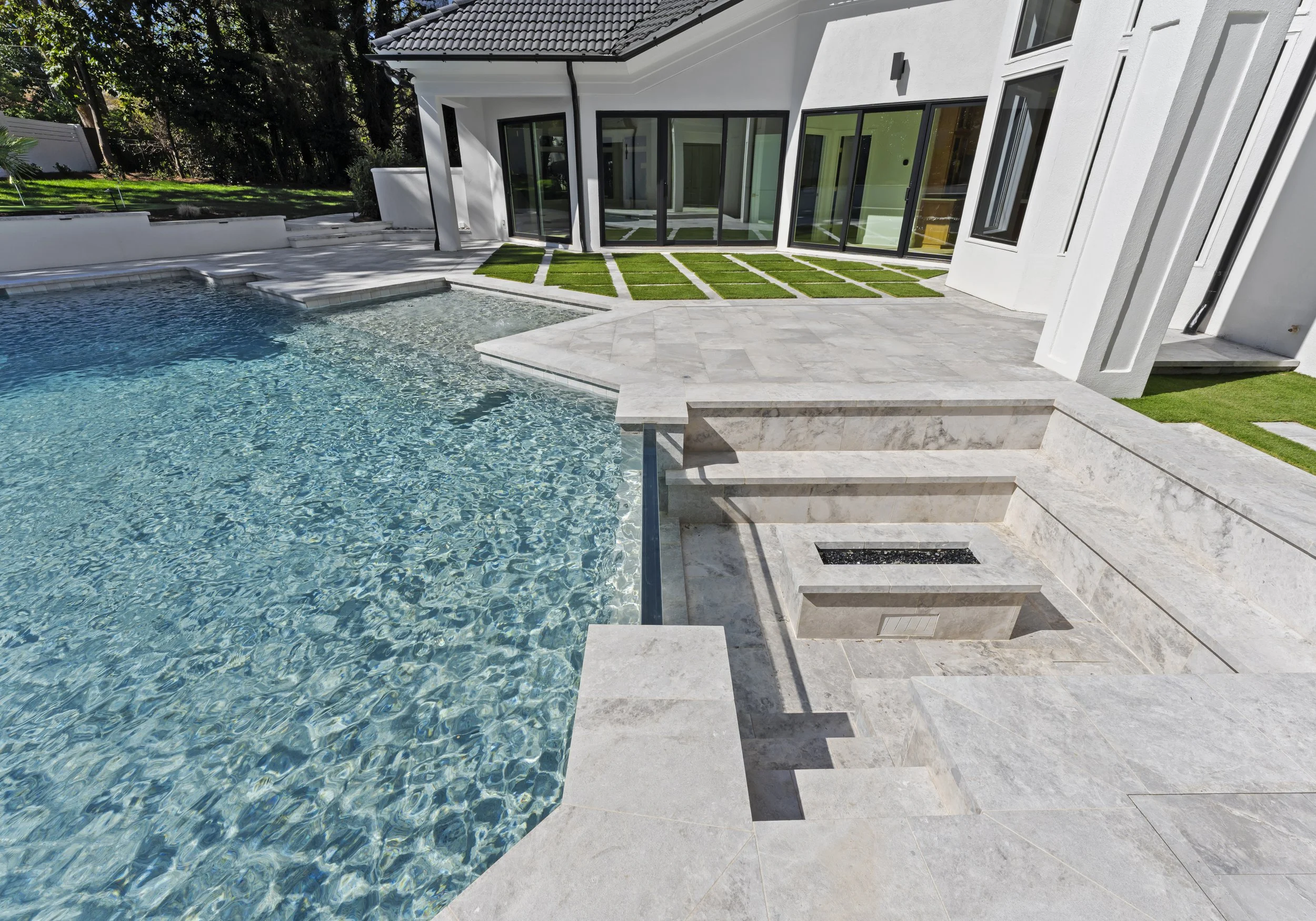 Modern house with a swimming pool in the backyard, featuring steps leading into the pool, a patio area with sections of artificial grass, large glass doors, and white exterior walls.