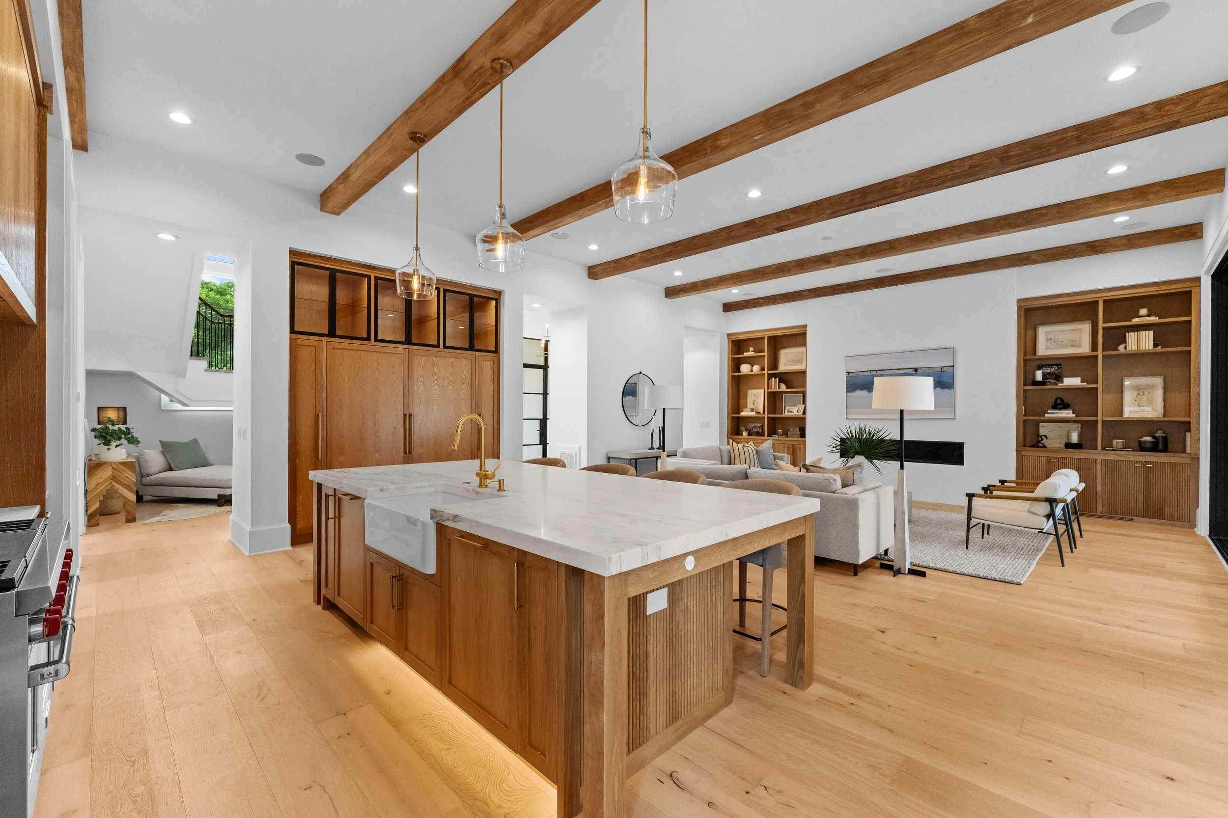 Open-concept living room with wooden beams, built-in shelves, a kitchen island with a marble countertop, and white furniture.