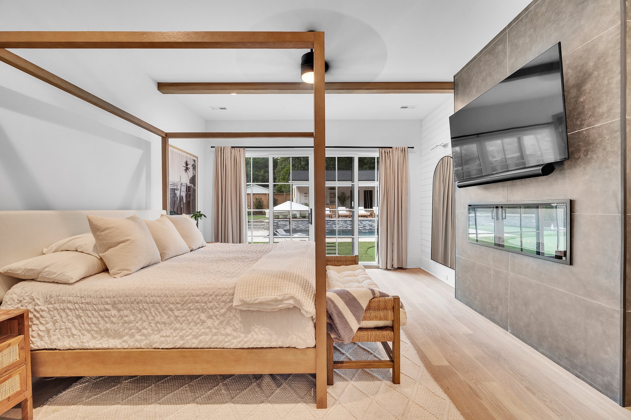 Modern bedroom with a wooden four-poster bed, cream bedding, a bedside table, and a large window with beige curtains overlooking a backyard with patio furniture.