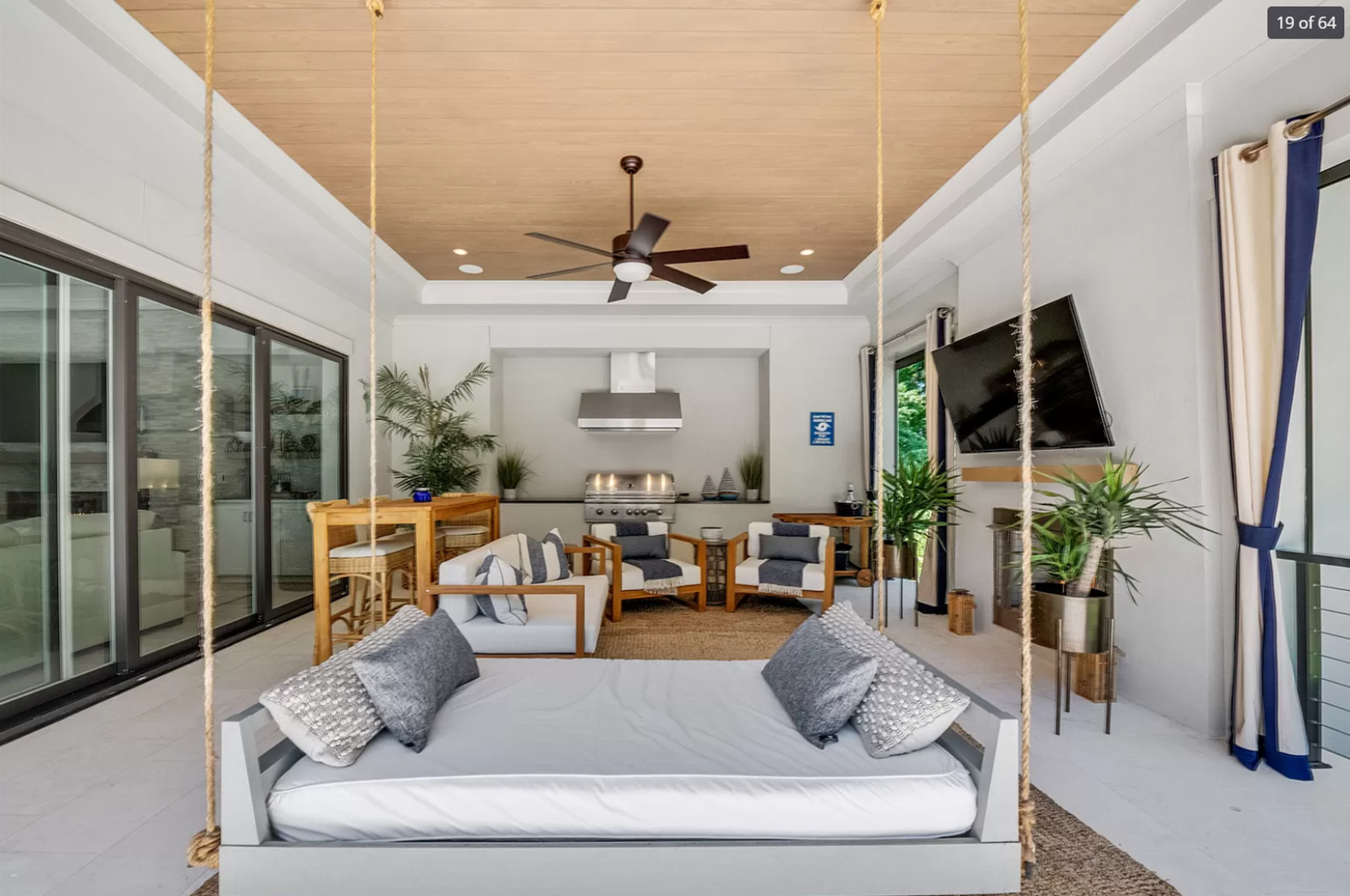 Coastal living room with a hanging bed, a seating area with white and gray pillows, a flat-screen TV, and sliding glass doors opening to a balcony with greenery.