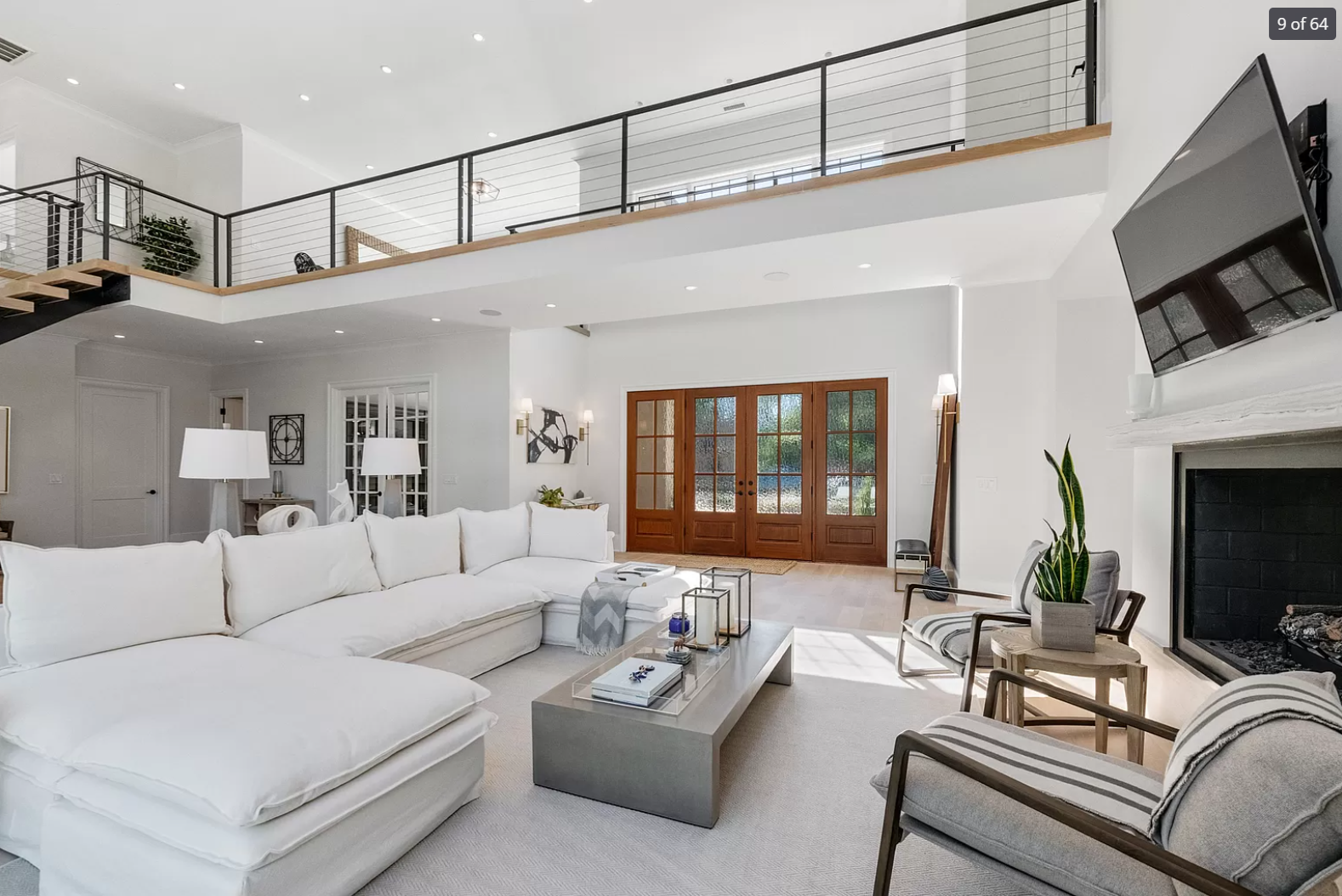 Bright living room with white sectional sofa, striped armchair, modern fireplace, wall-mounted TV, glass front door, and second-floor balcony with black railing.