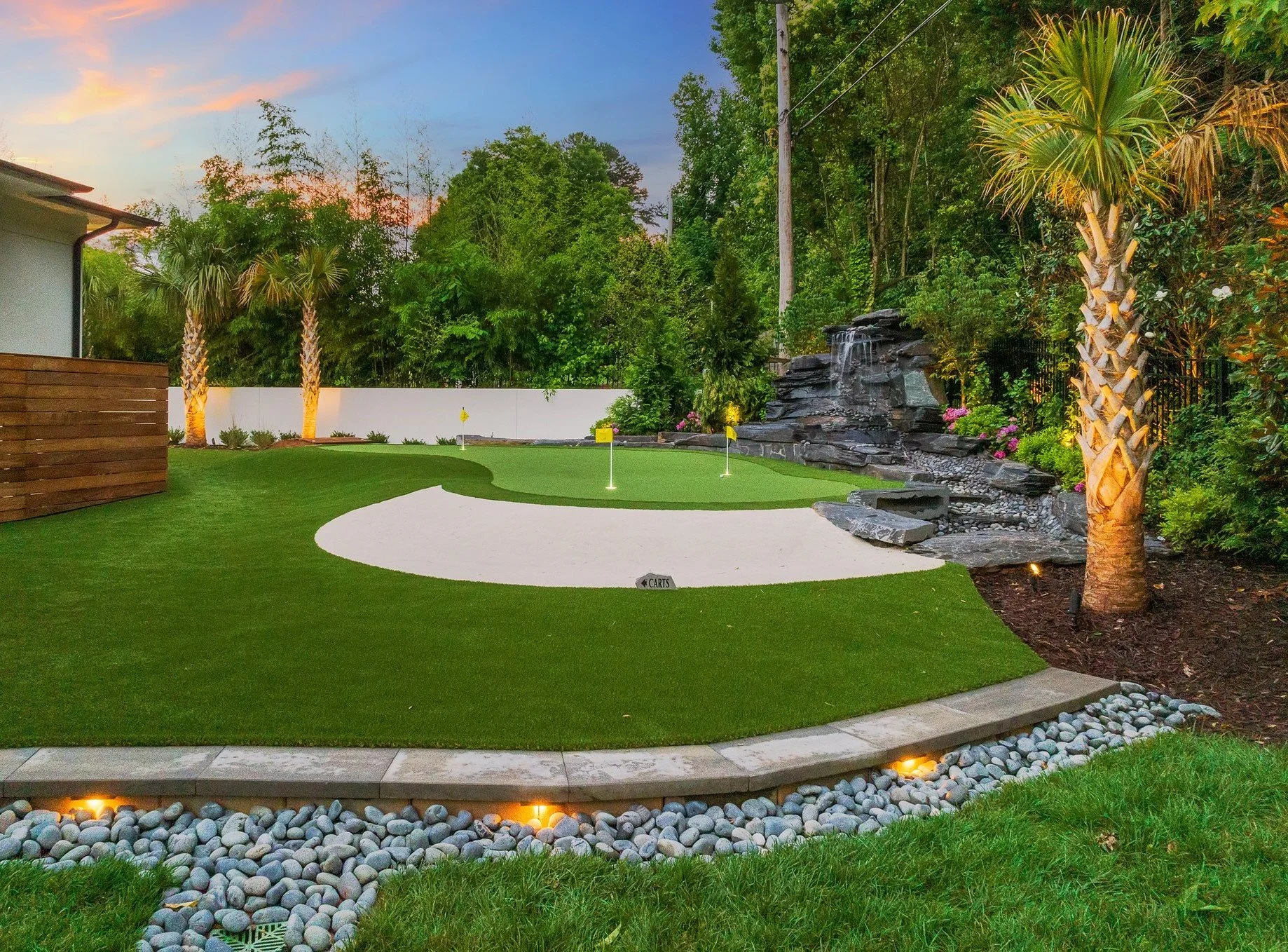 Backyard with a mini golf putting green, sand trap, water feature, palm trees, and surrounding landscaping.