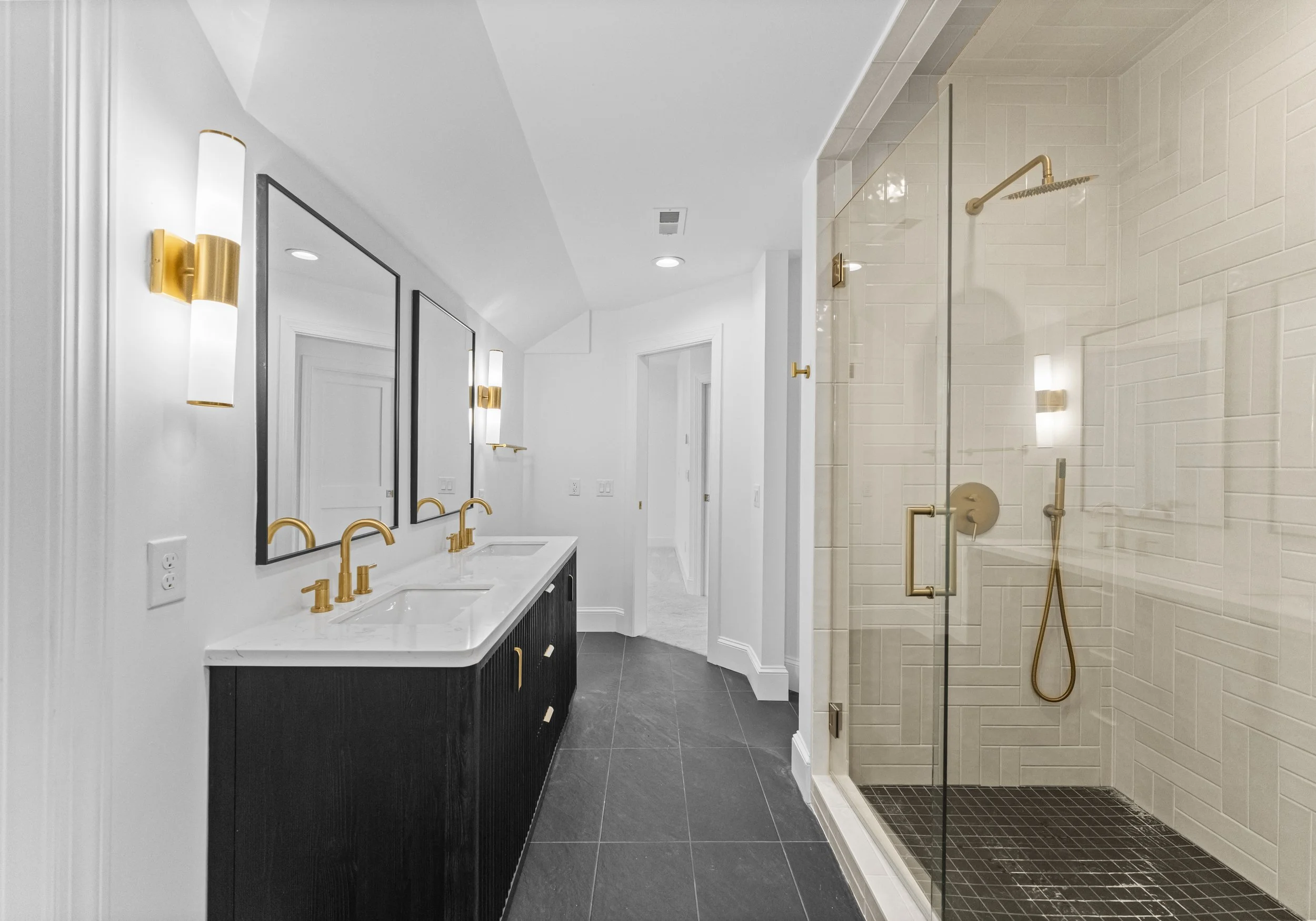 Modern bathroom with a double vanity with gold fixtures, black cabinets, and white marble countertop, large mirrors, wall-mounted sconces, and a glass-enclosed shower with matching gold fixtures, white tile interior, and black floor tiles.