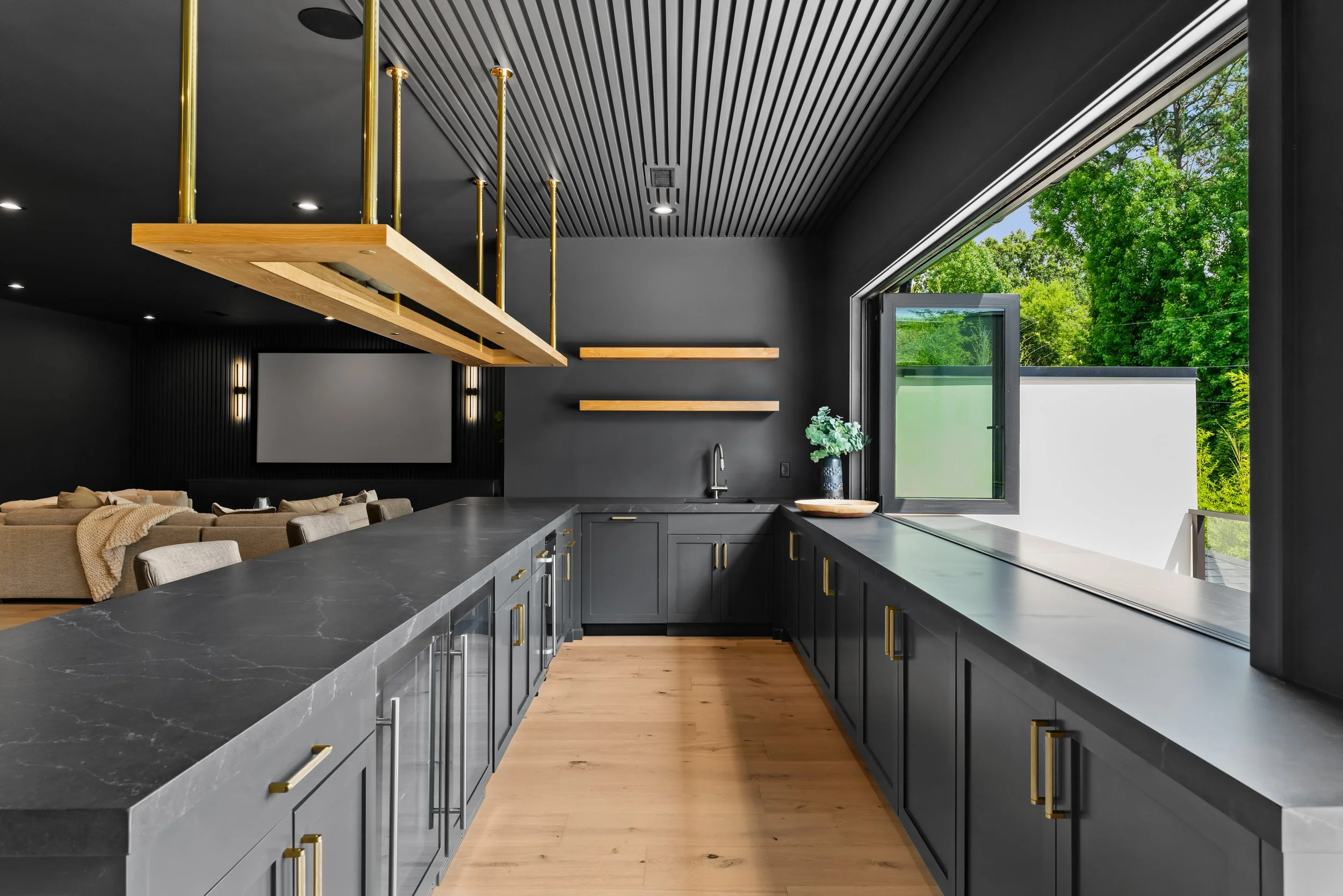 Modern kitchen with black cabinets, gray countertops, and a large open window overlooking green trees, with a living room with beige furniture in the background.