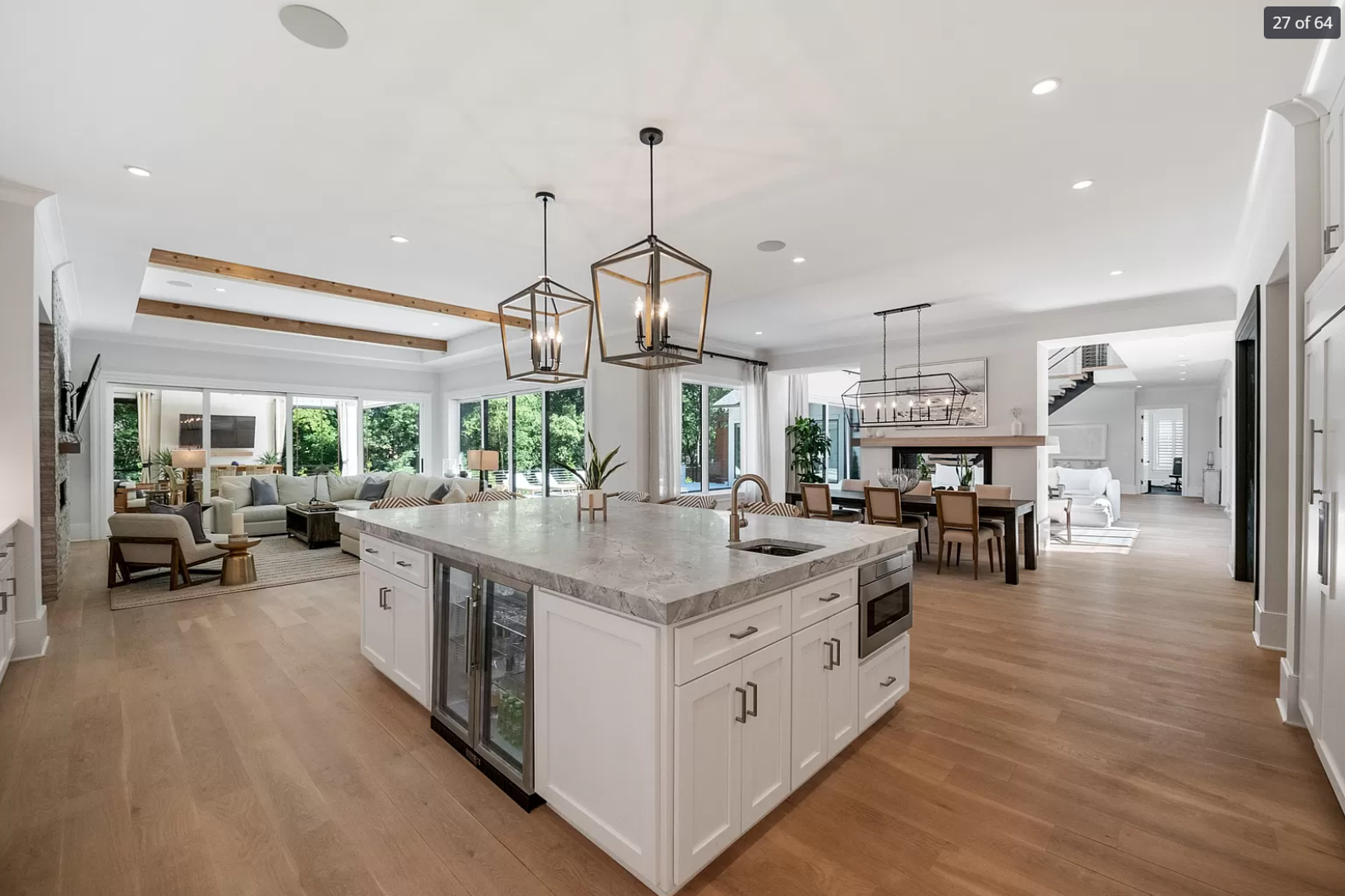 Open-concept living space with a kitchen island, dining area, and living room, featuring white walls, hardwood floors, and large windows with greenery outside.