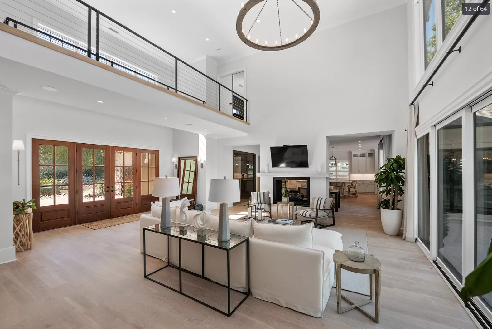 Spacious living room with white walls, large sliding glass doors, a fireplace, a wall-mounted TV, and modern furniture including a white sofa, striped armchairs, and side tables. A high ceiling with a round chandelier and an open upstairs balcony are